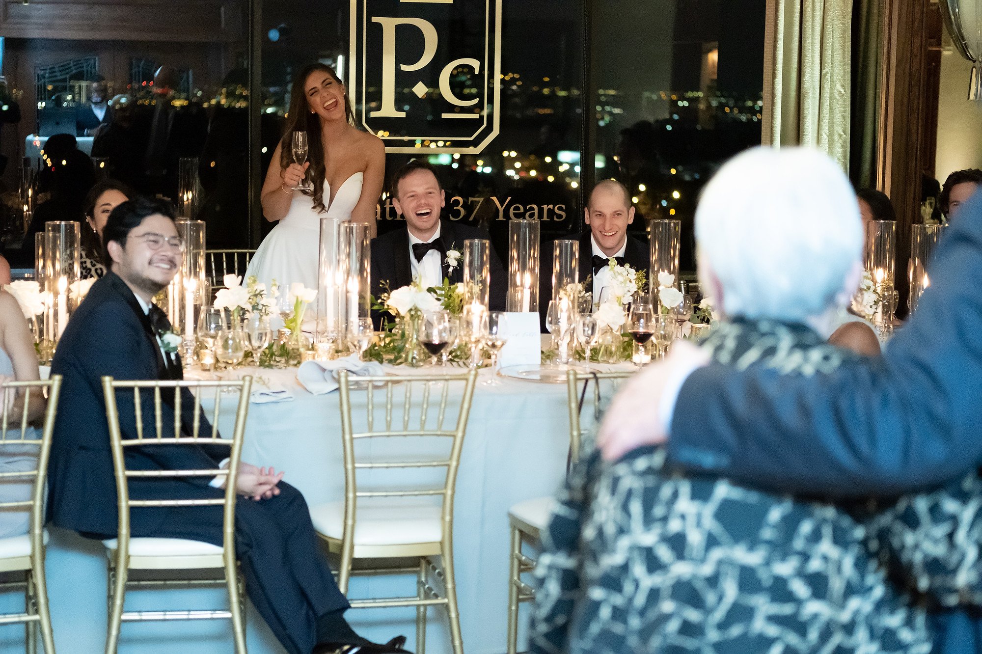 At a wedding reception, a bride in a white gown is standing and laughing, holding a glass of champagne. Seated at the table are men in tuxedos and women, all smiling and enjoying the moment. The table is decorated with flowers and candles, with a cit