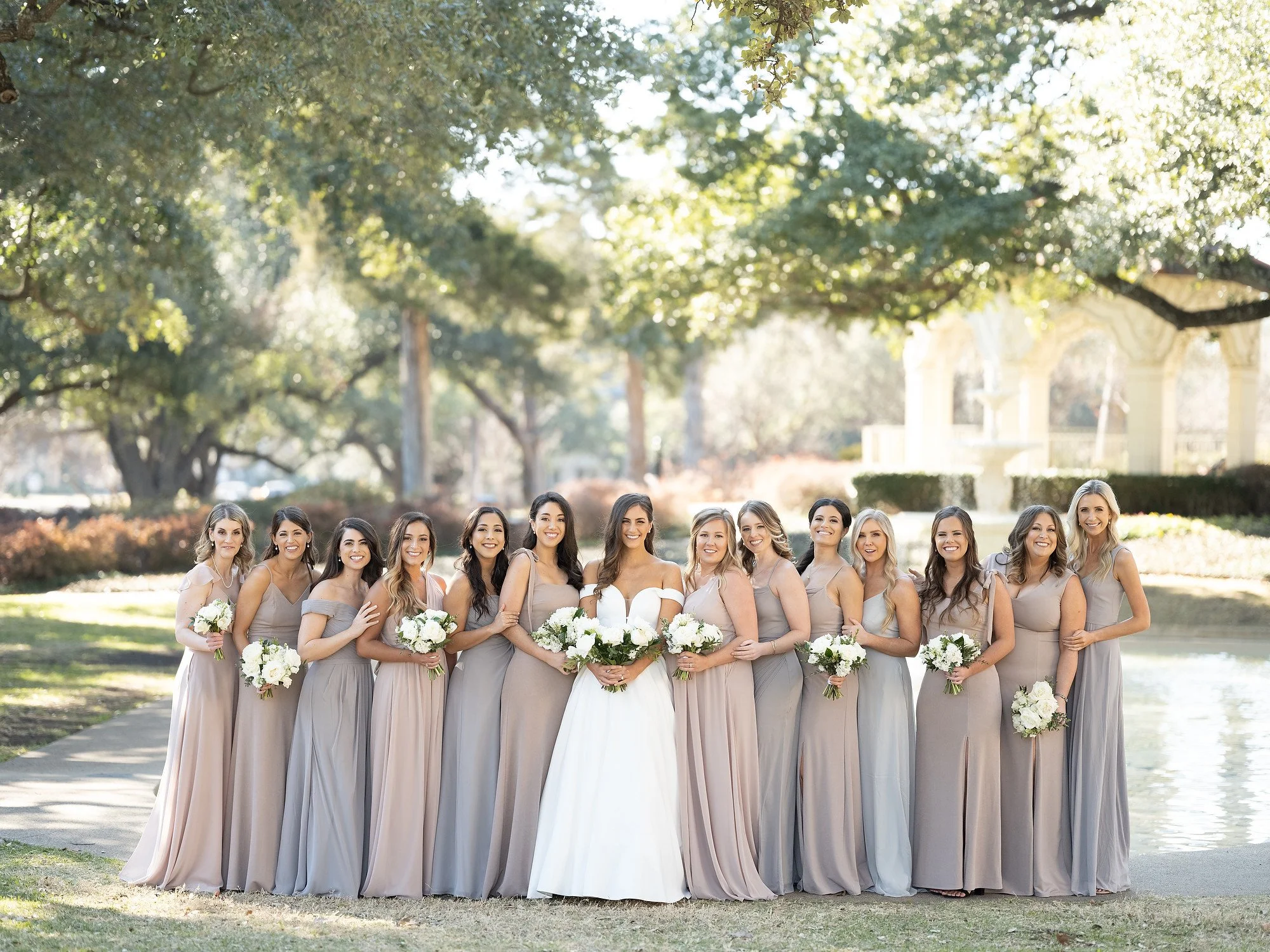 Bride and bridesmaids standing outdoors near water, holding bouquets of white flowers, dressed in light-colored gowns.