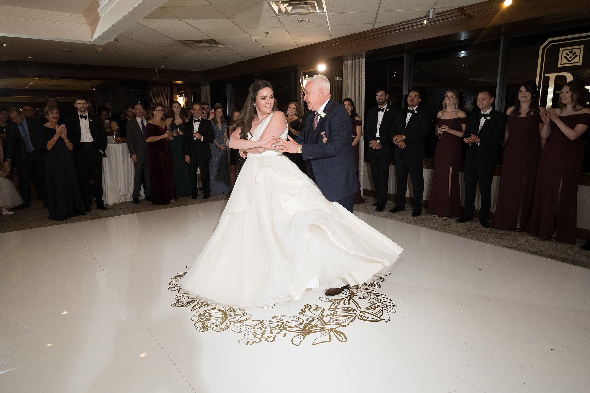 A bride in a white wedding gown is dancing with an older man in a navy suit on a decorated dance floor while guests in formal attire watch and applaud.
