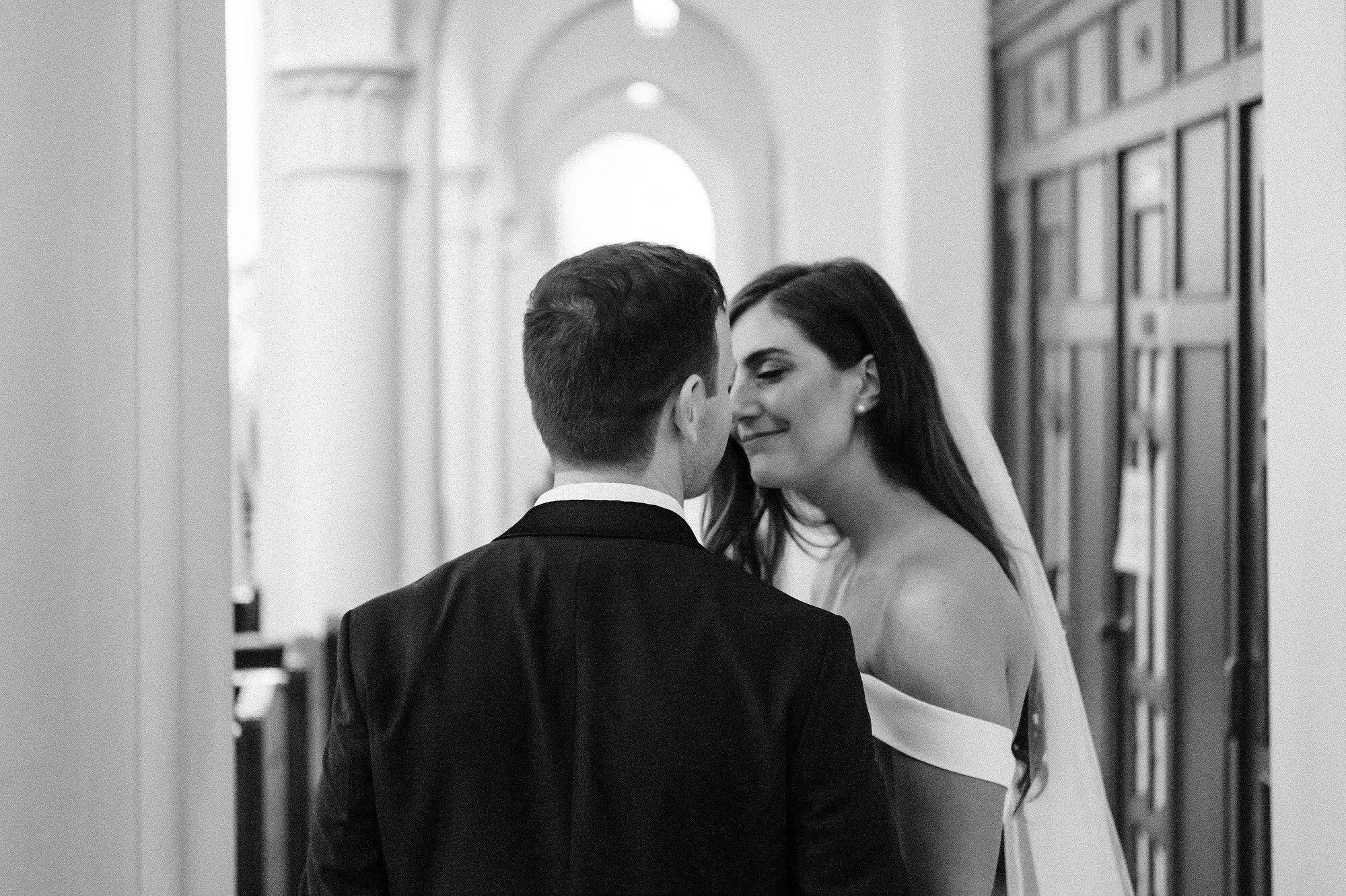A black and white photograph of a bride and groom in a hallway with ornate woodwork, standing close and smiling at each other.