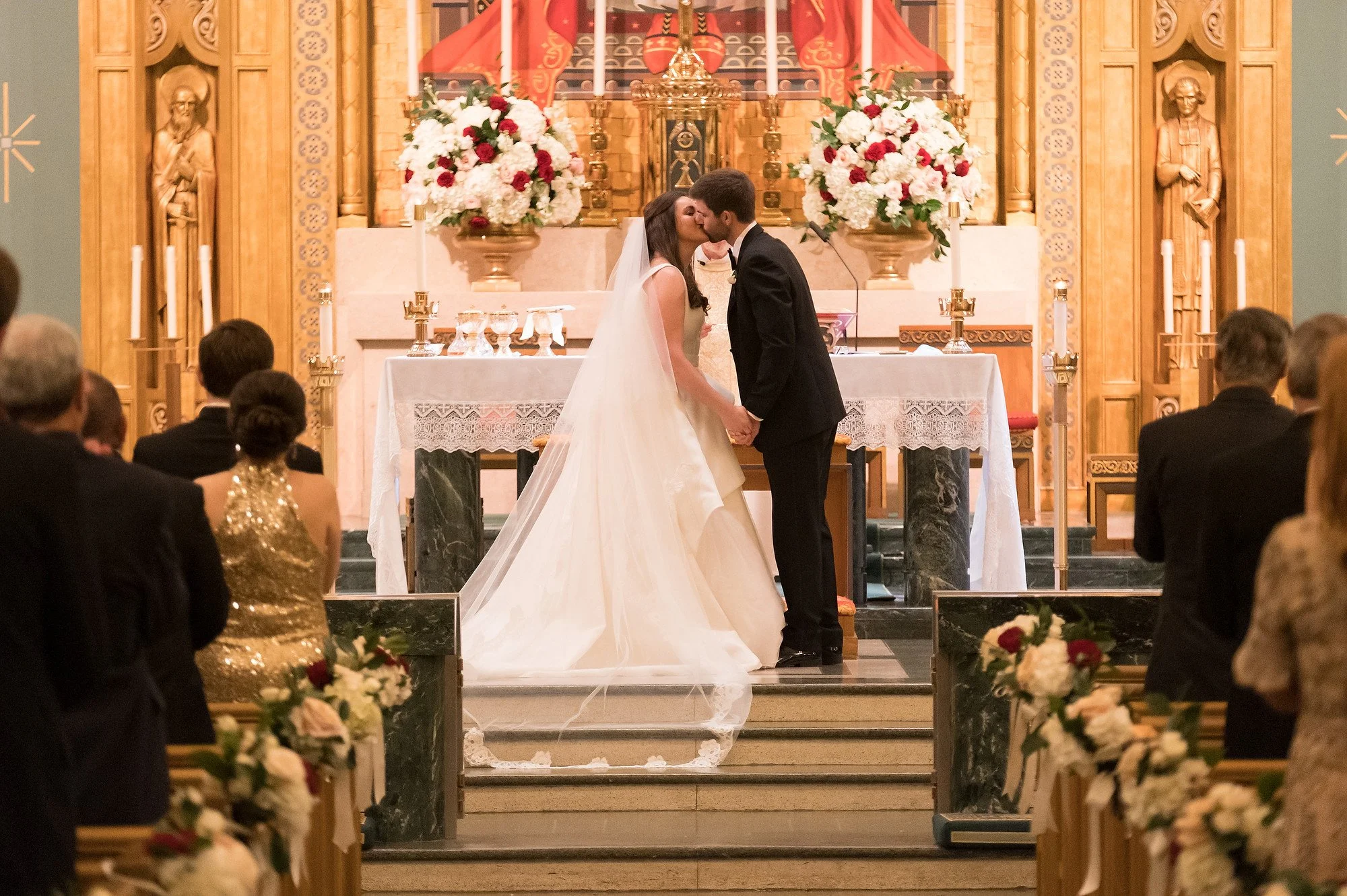 A bride and groom sharing their first kiss during a wedding ceremony in a church, with guests seated and floral arrangements in the background.