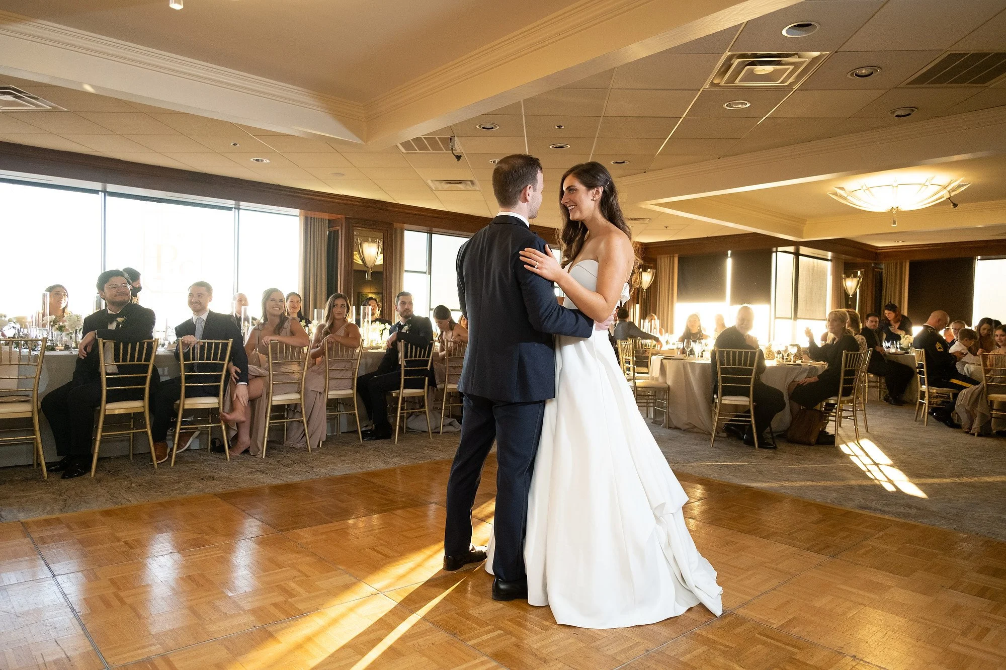 A bride and groom share a dance at their wedding reception, with guests seated at tables watching them in a well-lit banquet hall.