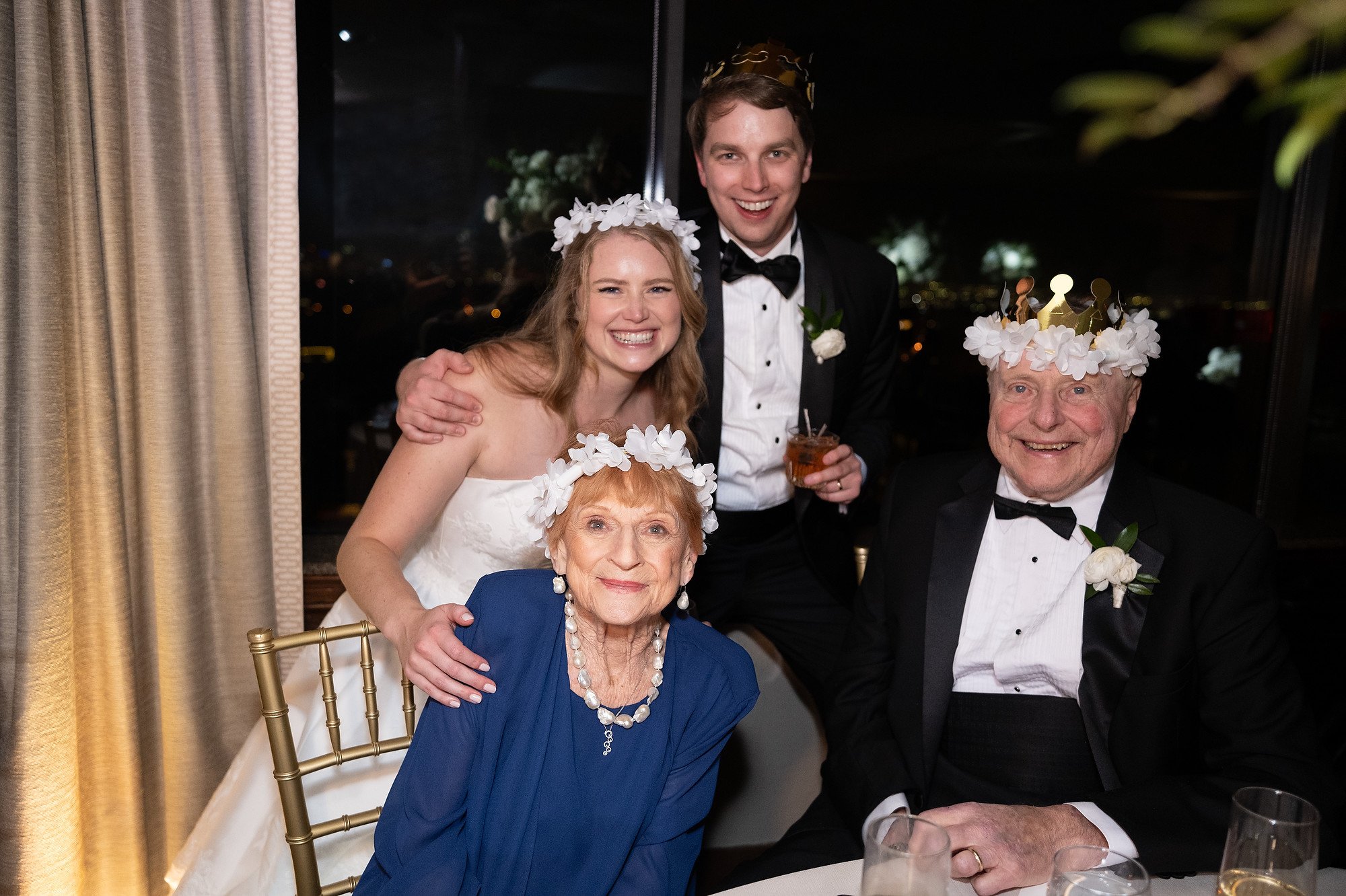 Four people at a wedding celebration, two women and two men, all wearing crowns and smiling. The woman in white dress and the man in tuxedo are likely the bride and groom, with the couple of older individuals possibly being family members. They are s