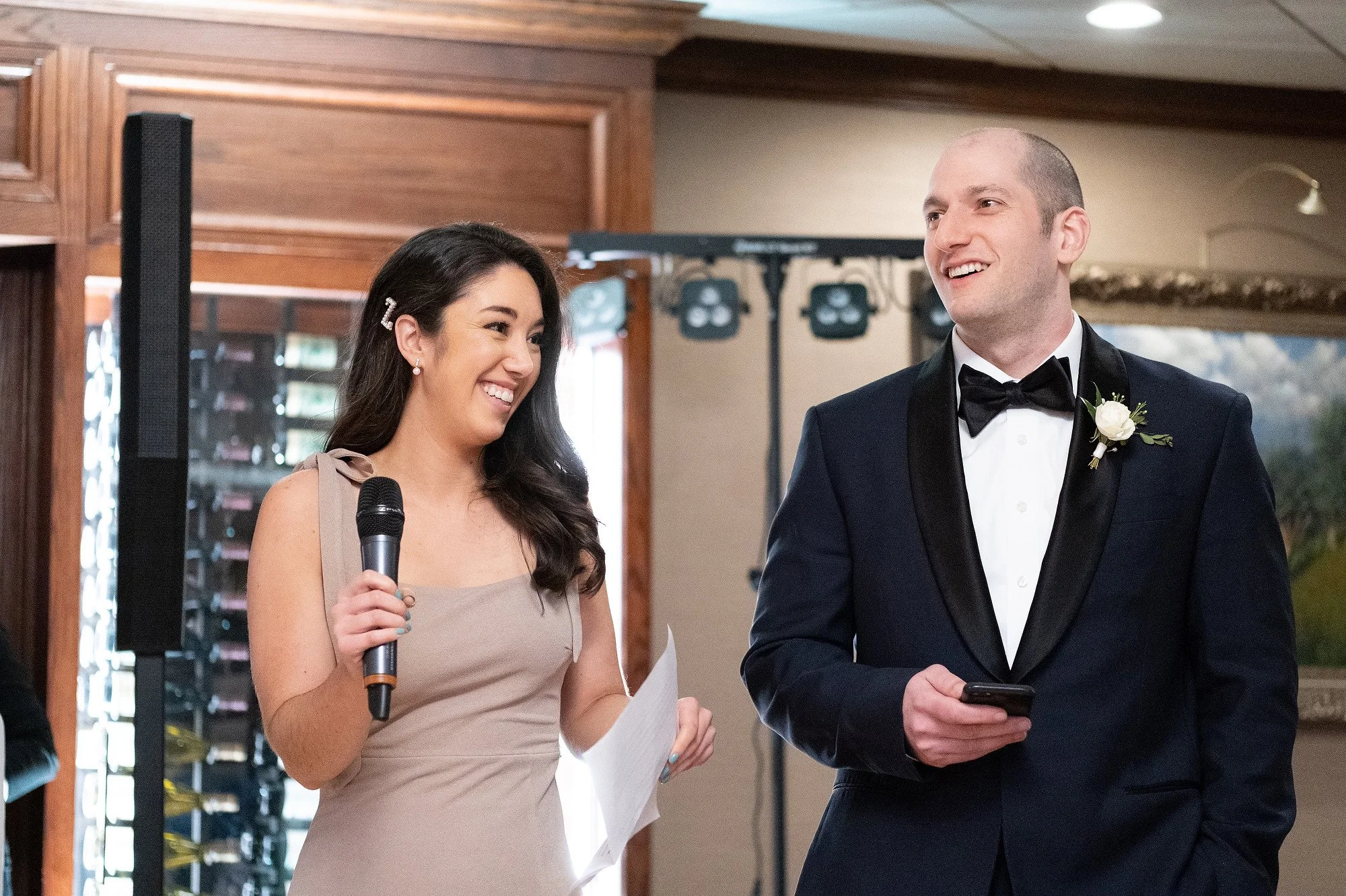 A woman in a beige dress holding a microphone and a piece of paper, smiling at a man in a tuxedo with a white boutonniere, who is holding a smartphone, at a wedding reception.