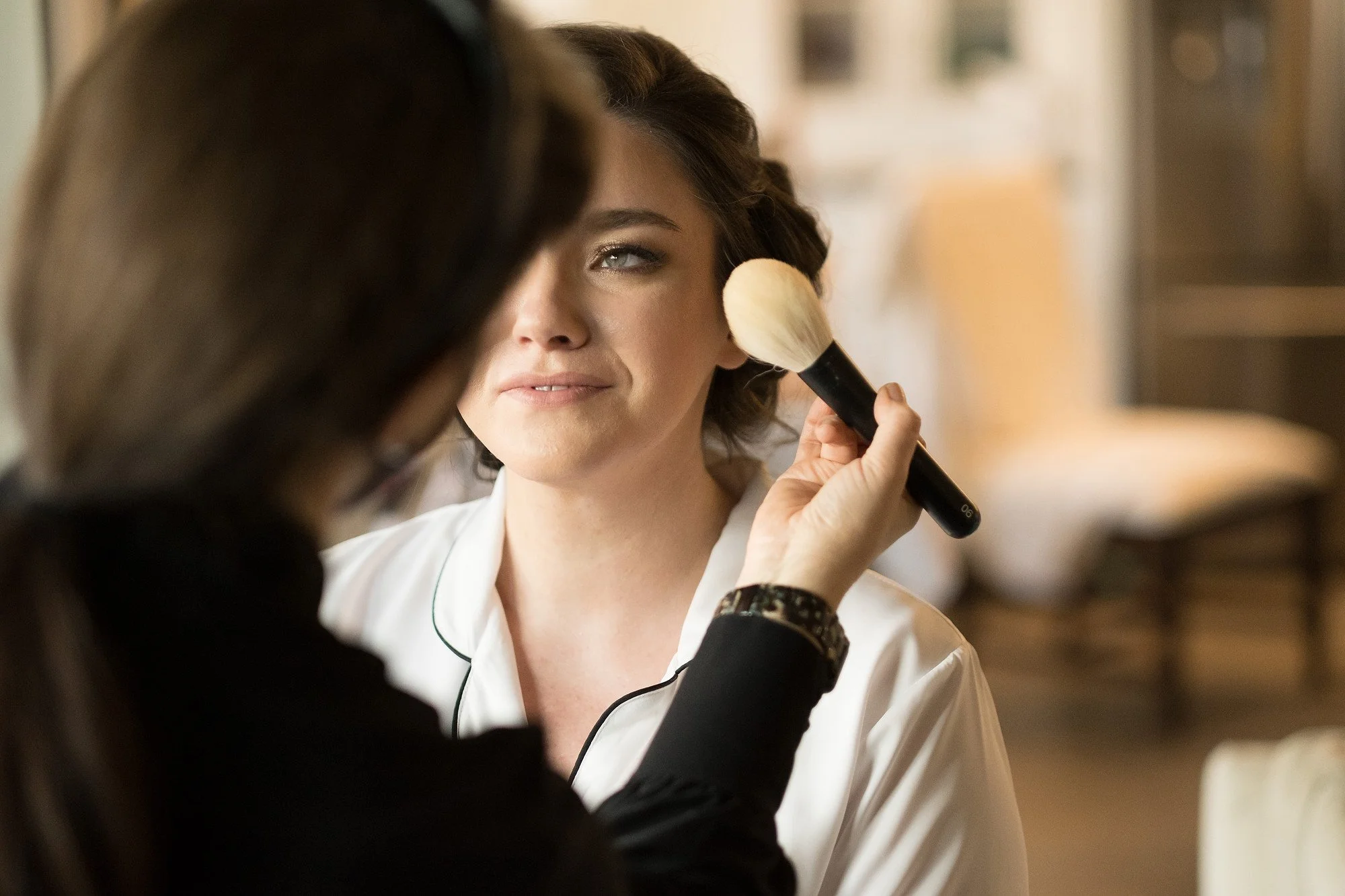 A woman with styled hair is getting her makeup done with a large brush by a makeup artist in a well-lit room.