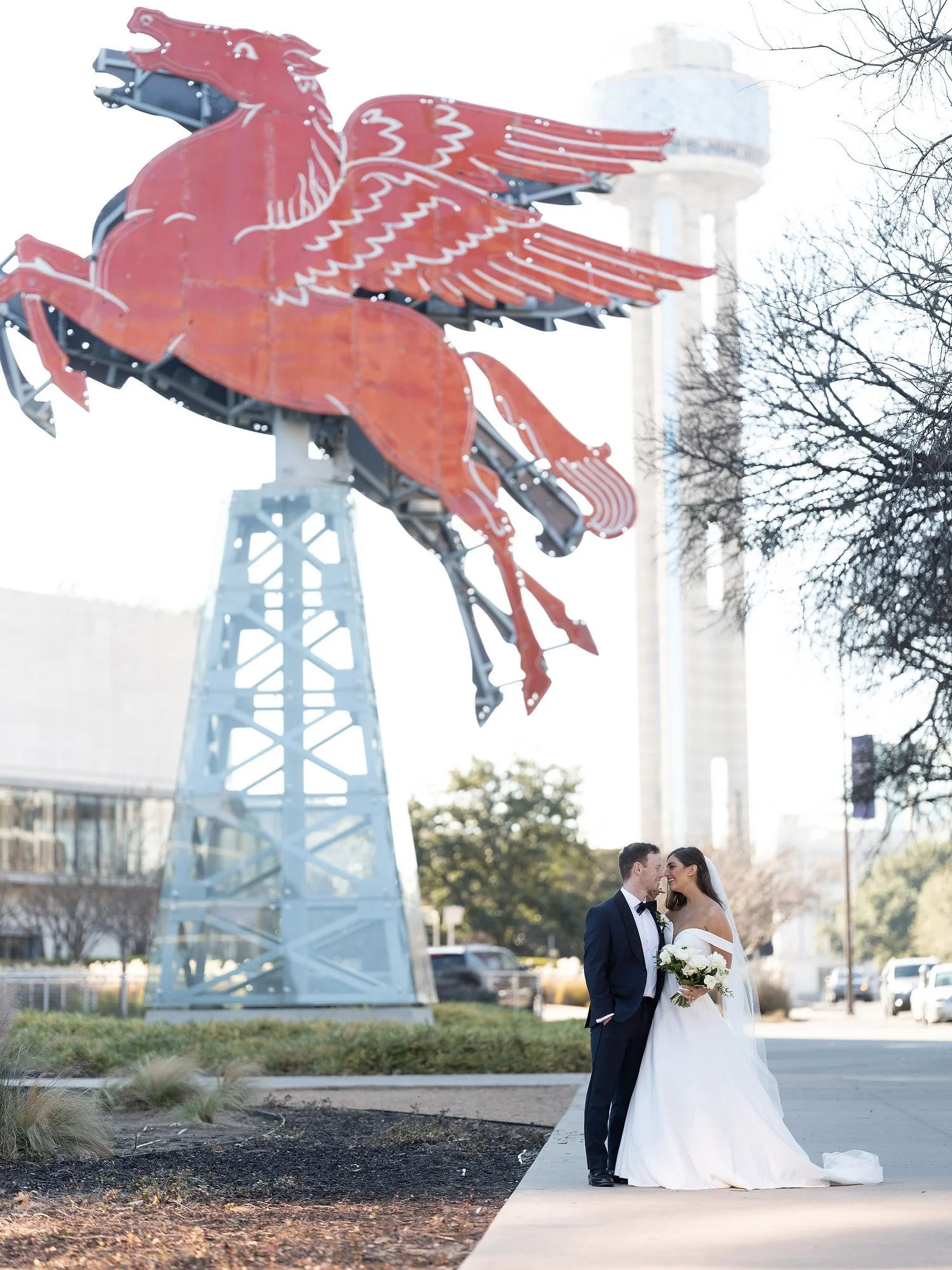A newlywed couple dressed in wedding attire standing outside near a large, red, neon-lit Pegasus sculpture with an urban cityscape in the background.
