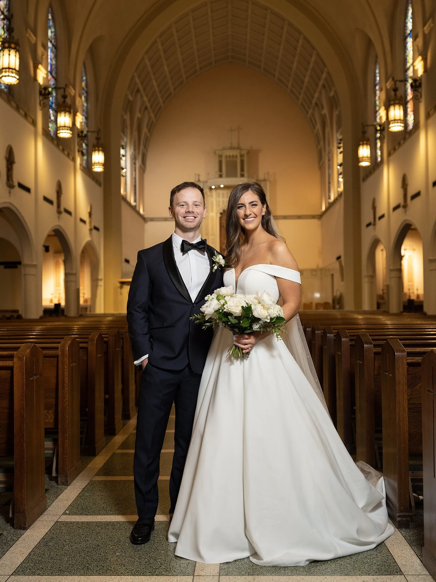 A bride and groom standing in a church, smiling for a wedding photo. The bride is wearing a white off-the-shoulder wedding gown and holding a bouquet of white roses, the groom is in a black tuxedo with a bow tie.