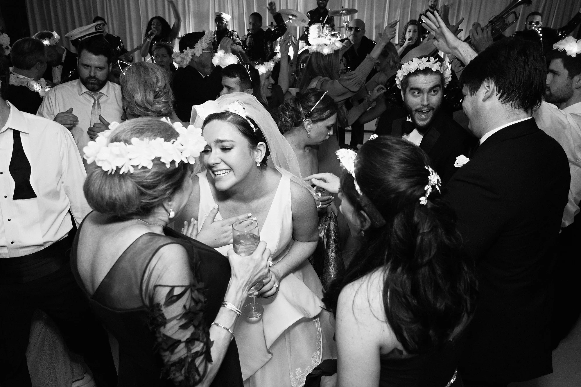 A wedding reception with guests dancing and celebrating, including a bride in a wedding dress and veil, surrounded by family and friends wearing floral crowns and formal attire.
