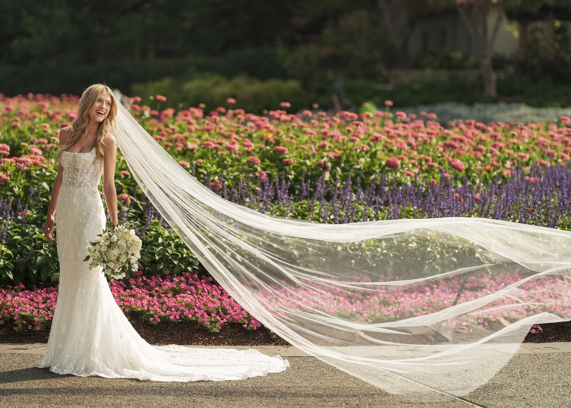 A smiling bride in a lace wedding gown with long veil holding a bouquet standing on a garden path with colorful flowers in the background.