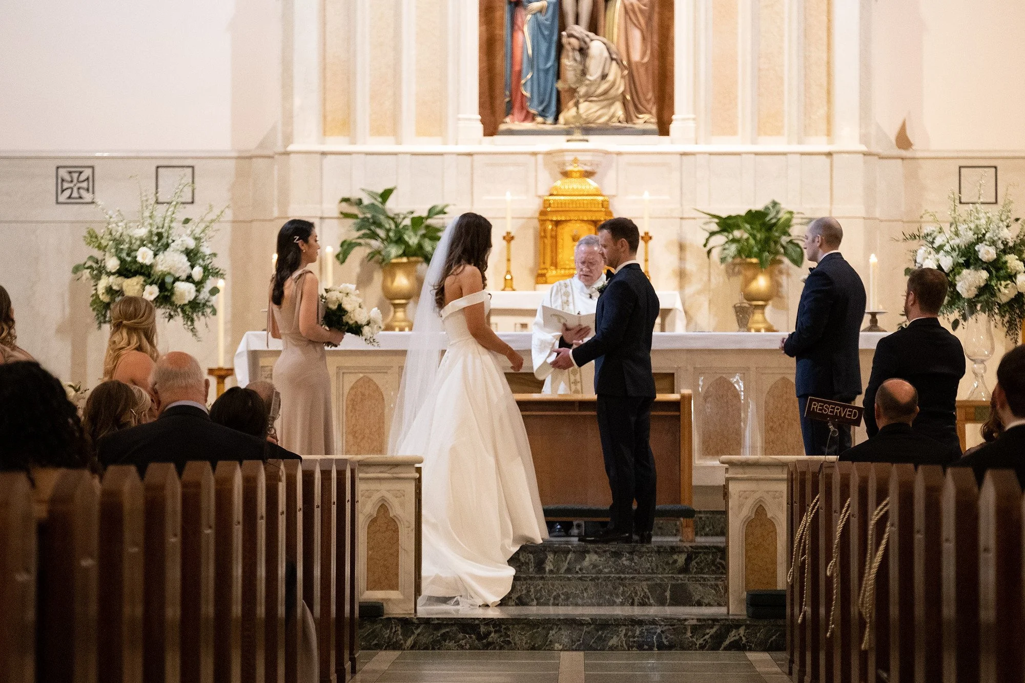 A couple getting married at an altar inside a church, with a priest officiating and a bridesmaid holding flowers, surrounded by wedding guests.