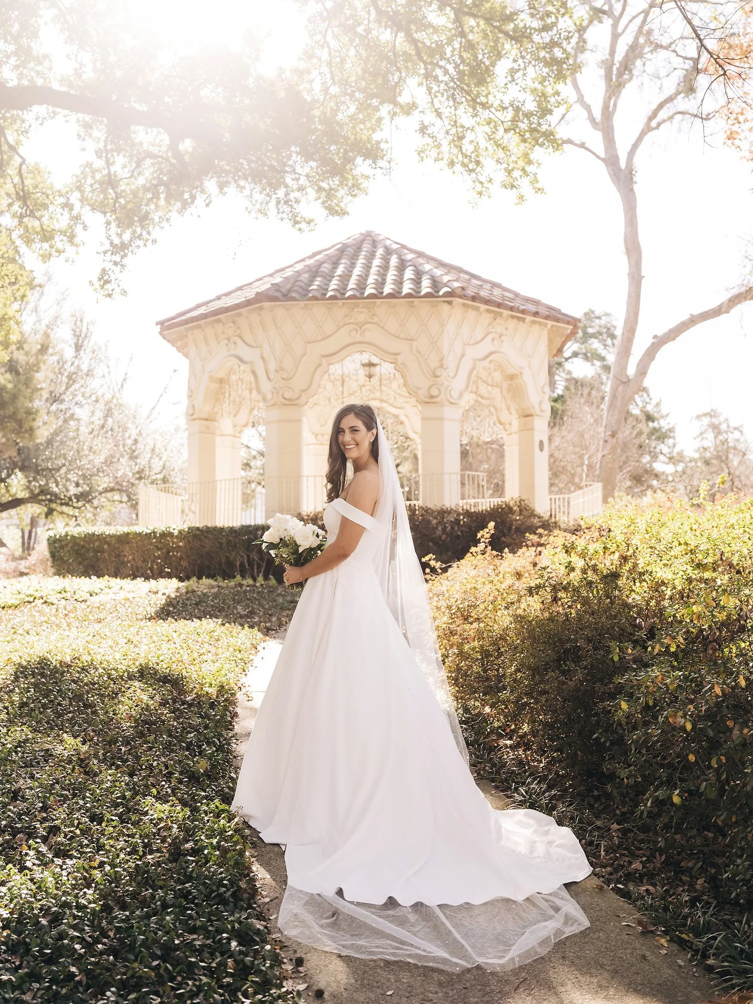 A bride in a white wedding gown holding a bouquet of white roses standing outdoors in front of a decorative gazebo, with sunlight filtering through trees.