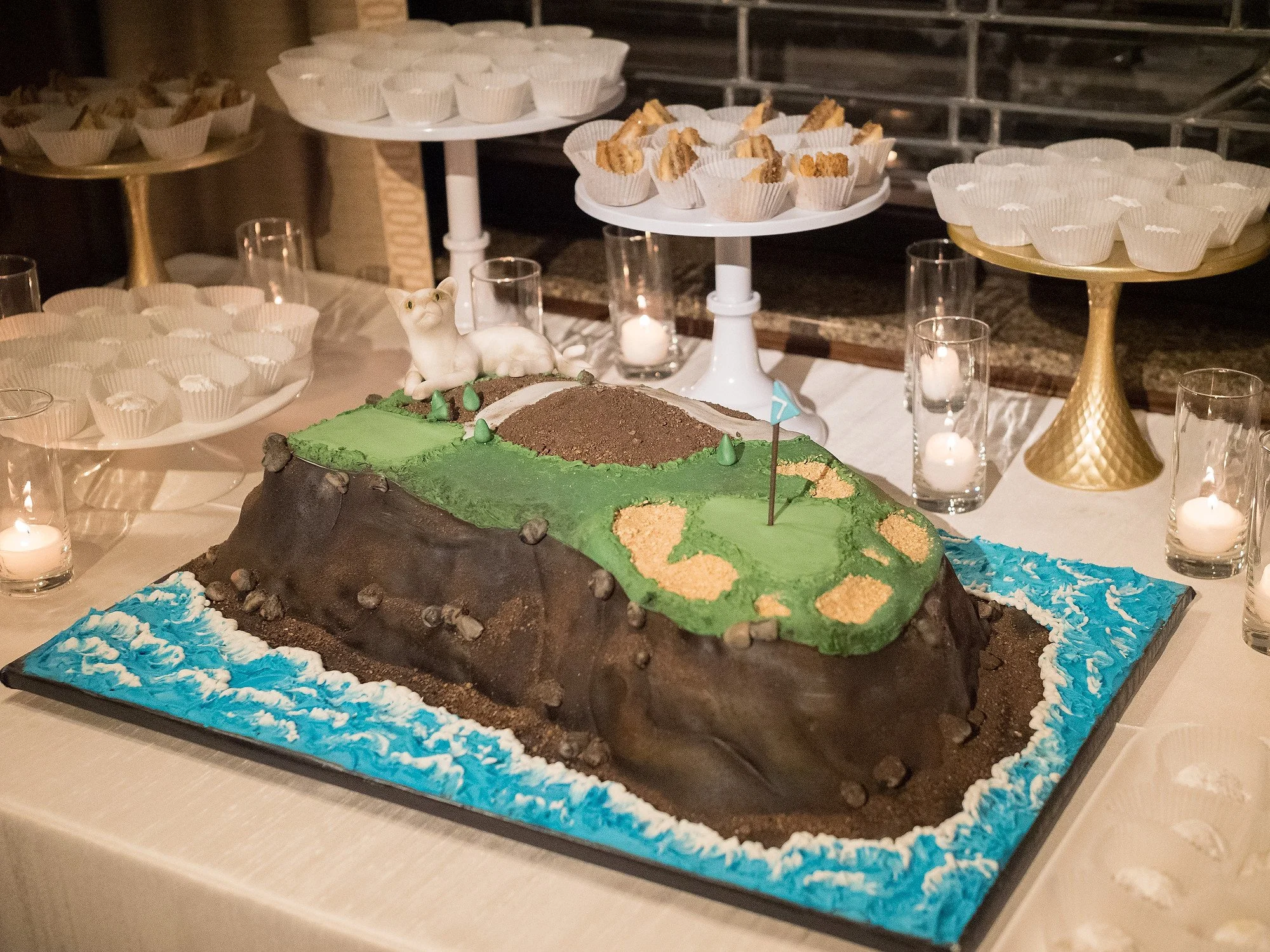 A cake shaped like a golf course with a mountain, green grass, a flag, a white animal figure, and tiny trees, surrounded by cupcakes and candles.