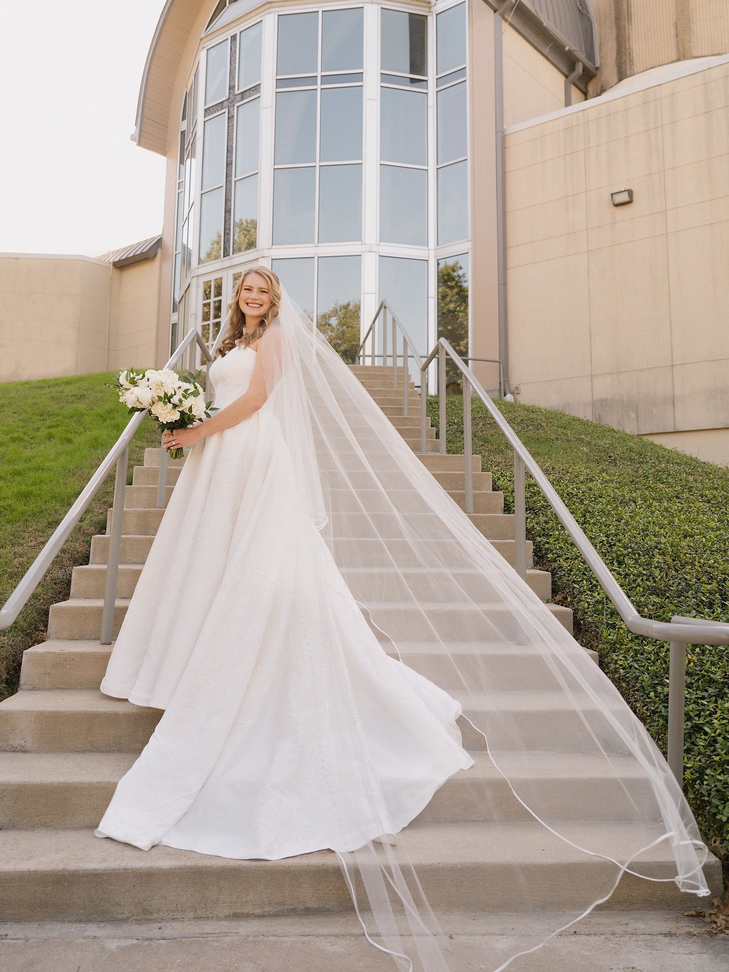A bride in a white wedding gown with a long train and veil, holding a bouquet of white flowers, standing on outdoor steps in front of a modern building with large glass windows.