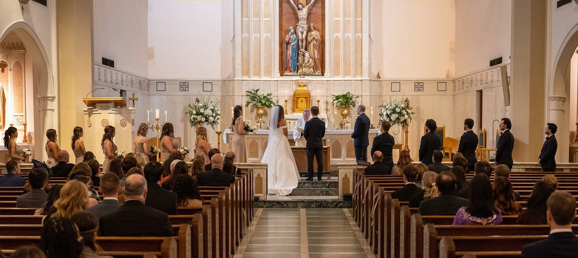 A wedding ceremony inside a church with the bride and groom standing at the altar, surrounded by bridesmaids and groomsmen, with guests seated in pews.