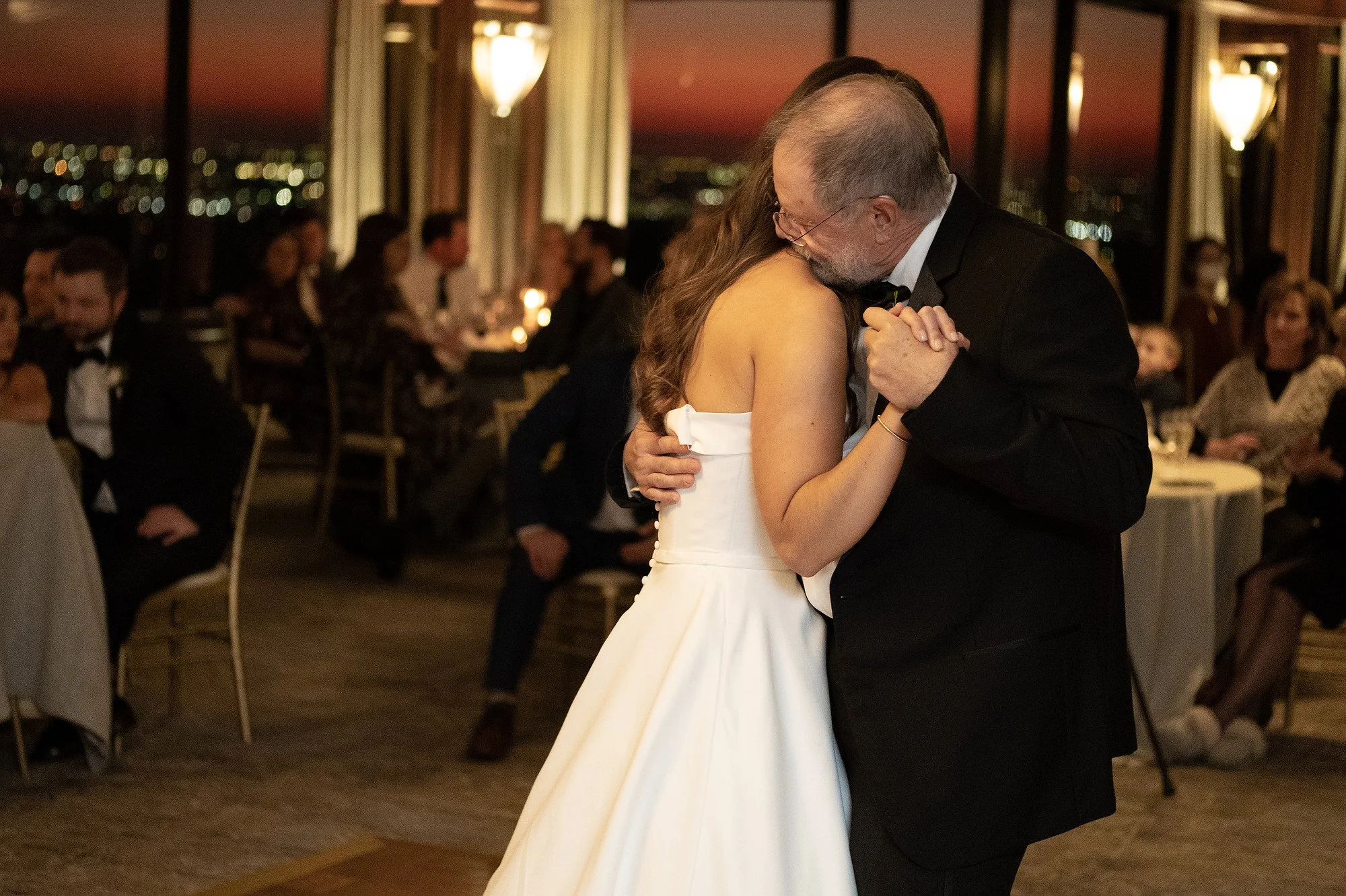 A bride and an older man, possibly her father, sharing a dance at a wedding reception in a dimly lit room with large windows and city lights outside.