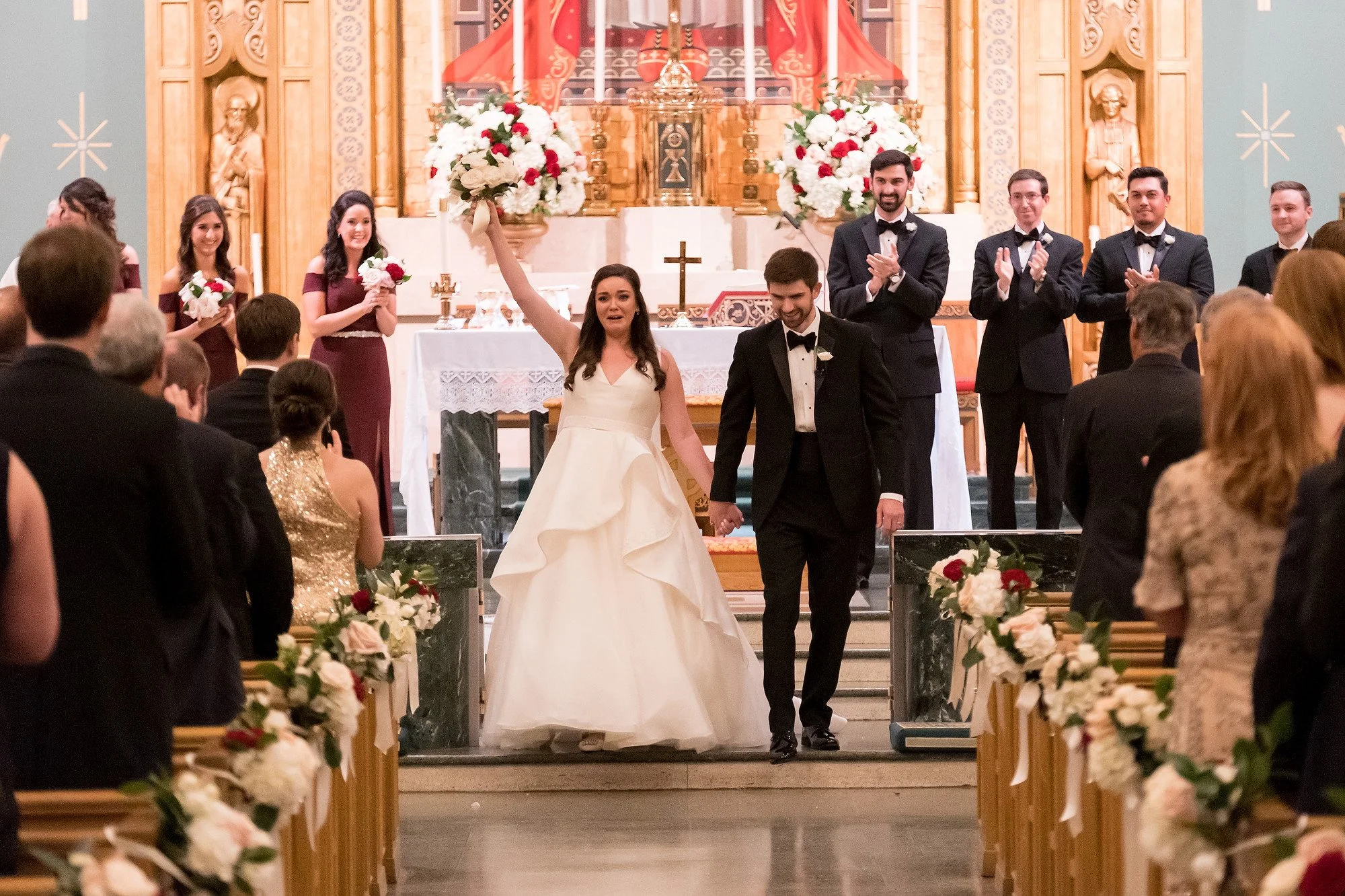 A bride and groom celebrating their wedding in a church, holding hands and smiling, with the bride raising a bouquet of flowers in the air. Bridesmaids and groomsmen stand behind them, clapping, as the congregation looks on. The church is decorated w