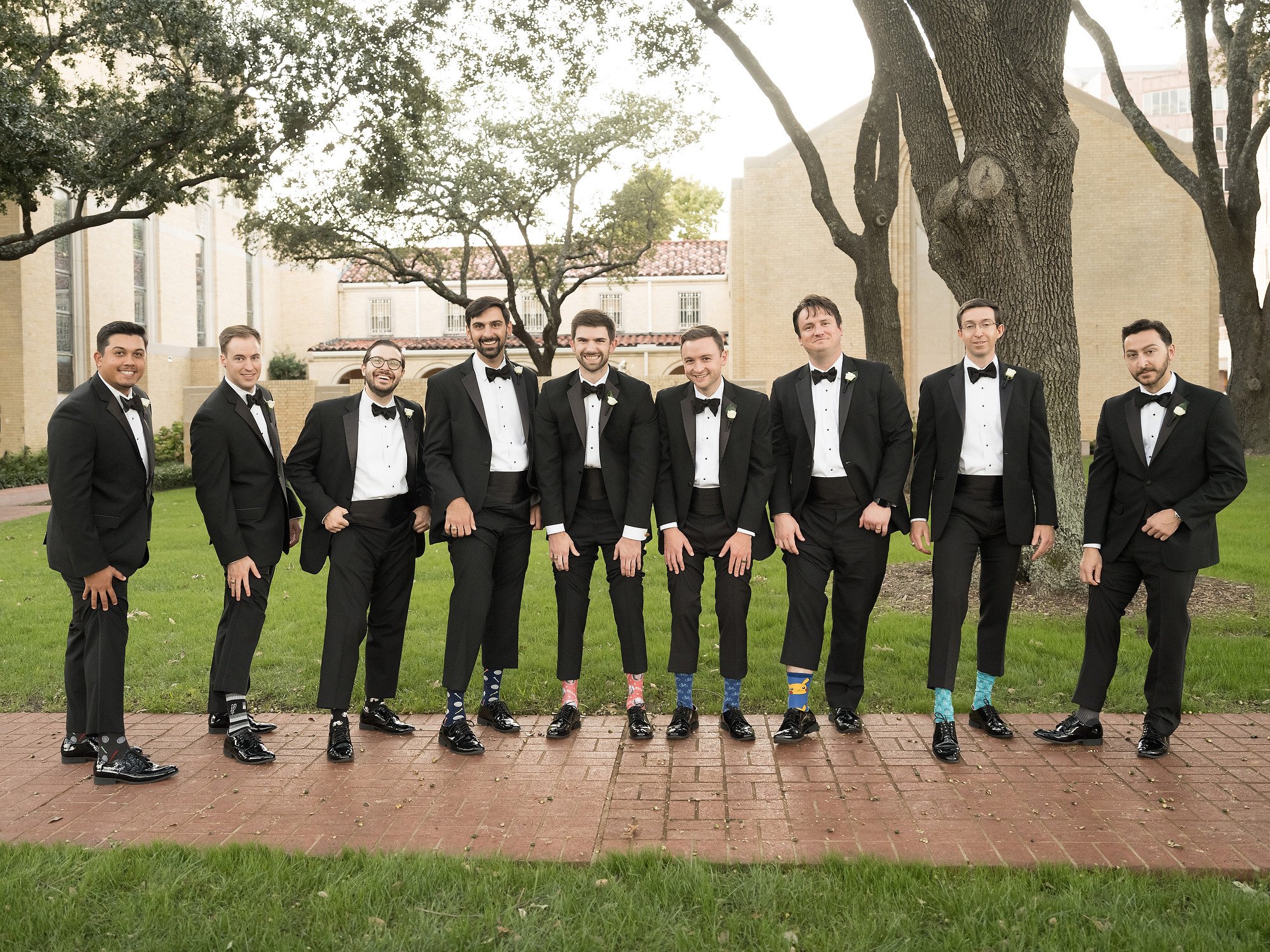 Group of men dressed in tuxedos with bow ties, standing outdoors on a brick pathway, posing for a photo.
