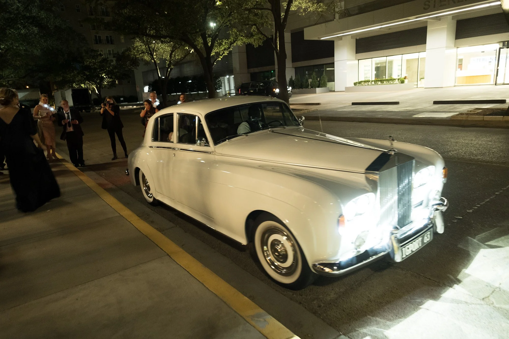 Vintage white luxury car with bright headlights, parked on a city street at night, with people dressed formally and taking photos nearby.