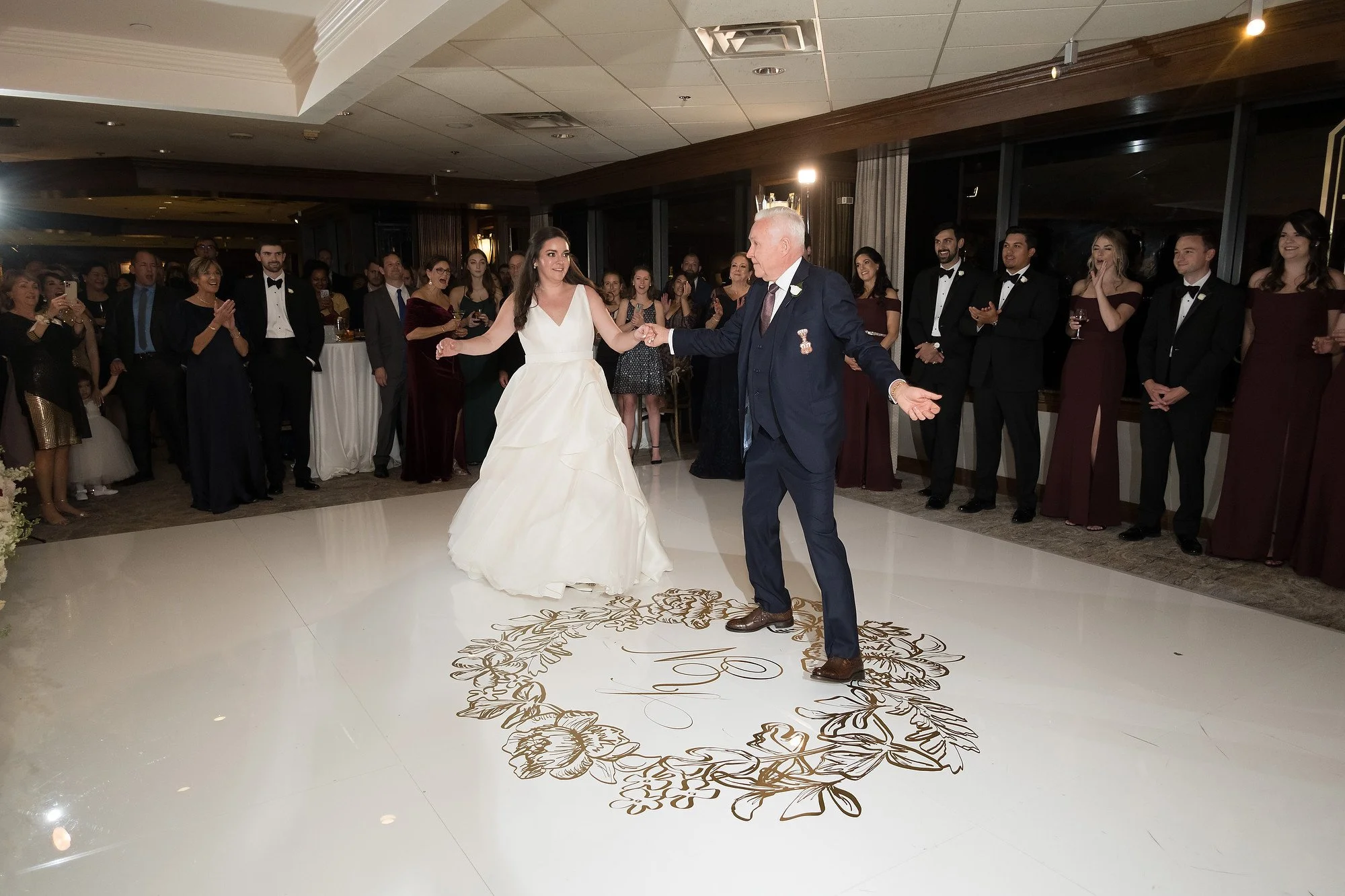 A bride and an older man dancing together on a wedding dance floor, surrounded by guests in formal attire, with some clapping and smiling, at a wedding reception.