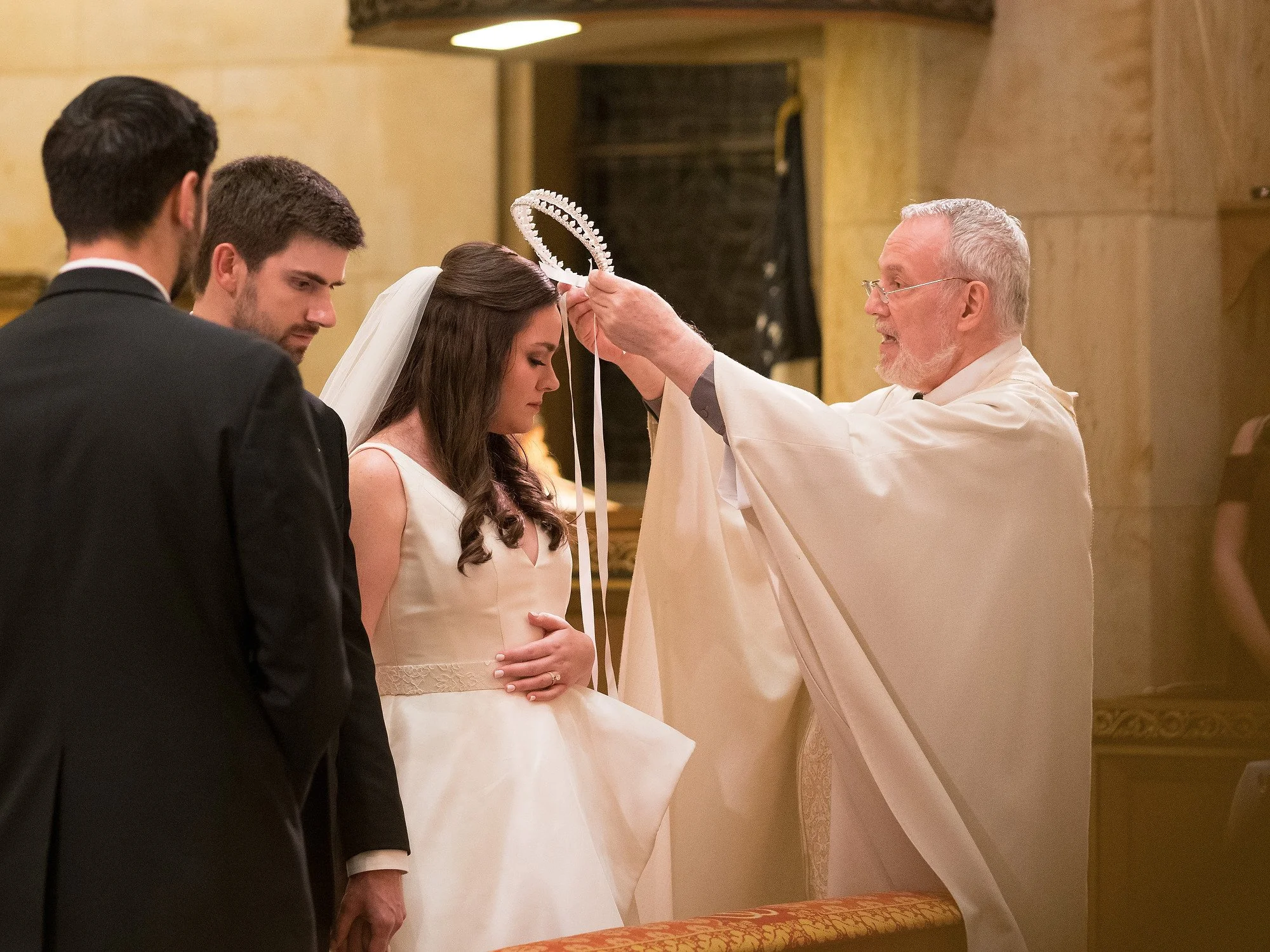A priest placing a tiara on a bride during a wedding ceremony, with two grooms standing beside her.