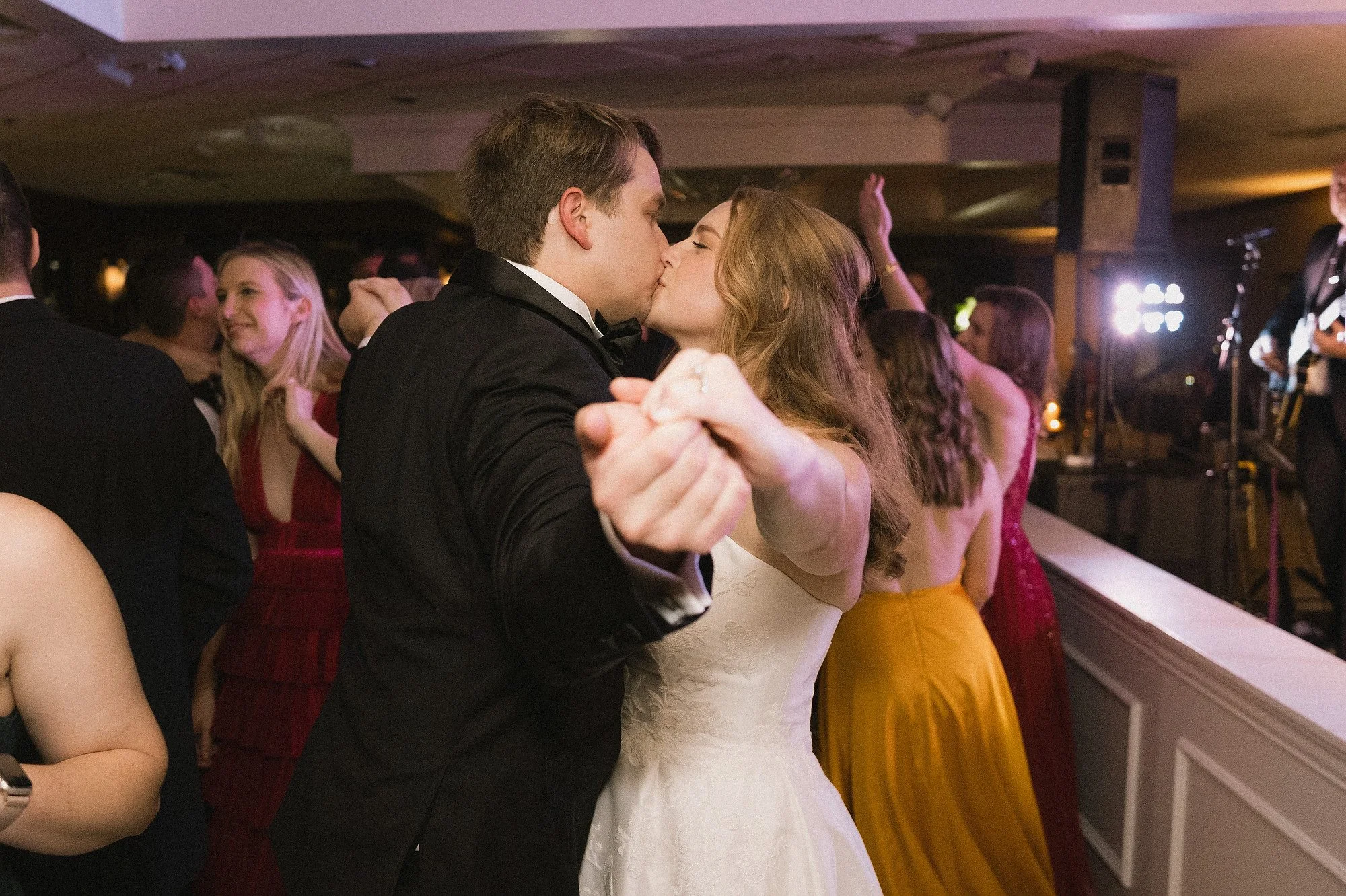 A couple in formal attire sharing a kiss while dancing at a wedding reception with other guests and a band in the background.