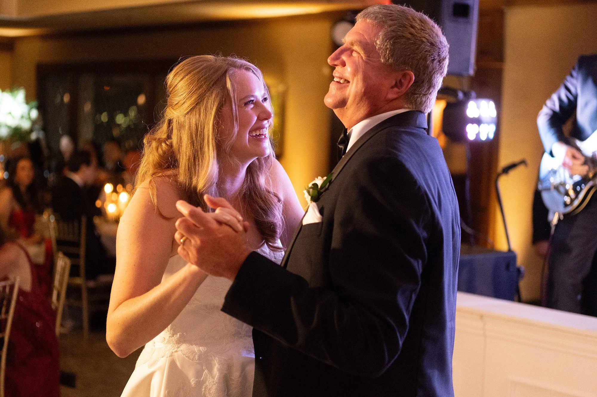 A bride and an older man, likely her father, dancing together at a wedding reception, smiling and holding hands, with a band playing in the background.