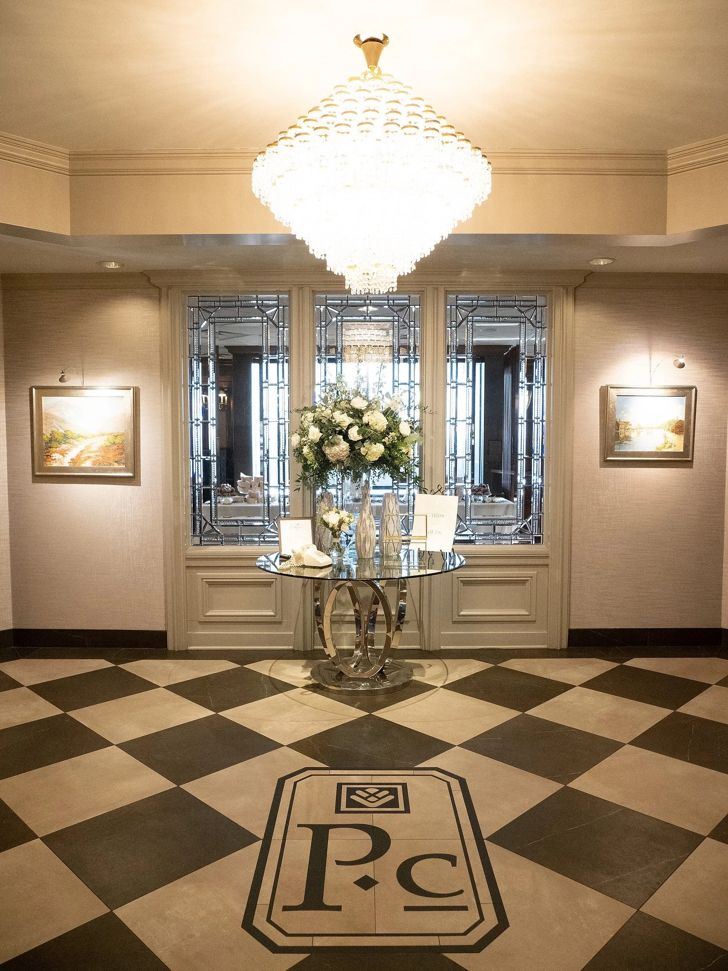 Elegant hotel or event lobby with a large chandelier, a glass table with a floral centerpiece, and black and white checkered flooring with a 'P C' monogram.