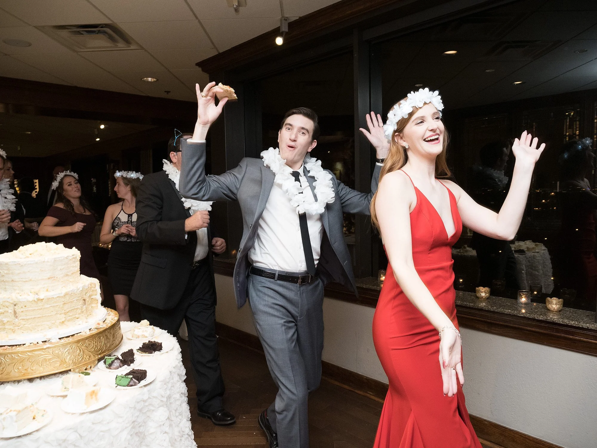 People celebrating at a wedding reception, with a woman in a red dress and a man in a gray suit dancing near a table with cake.