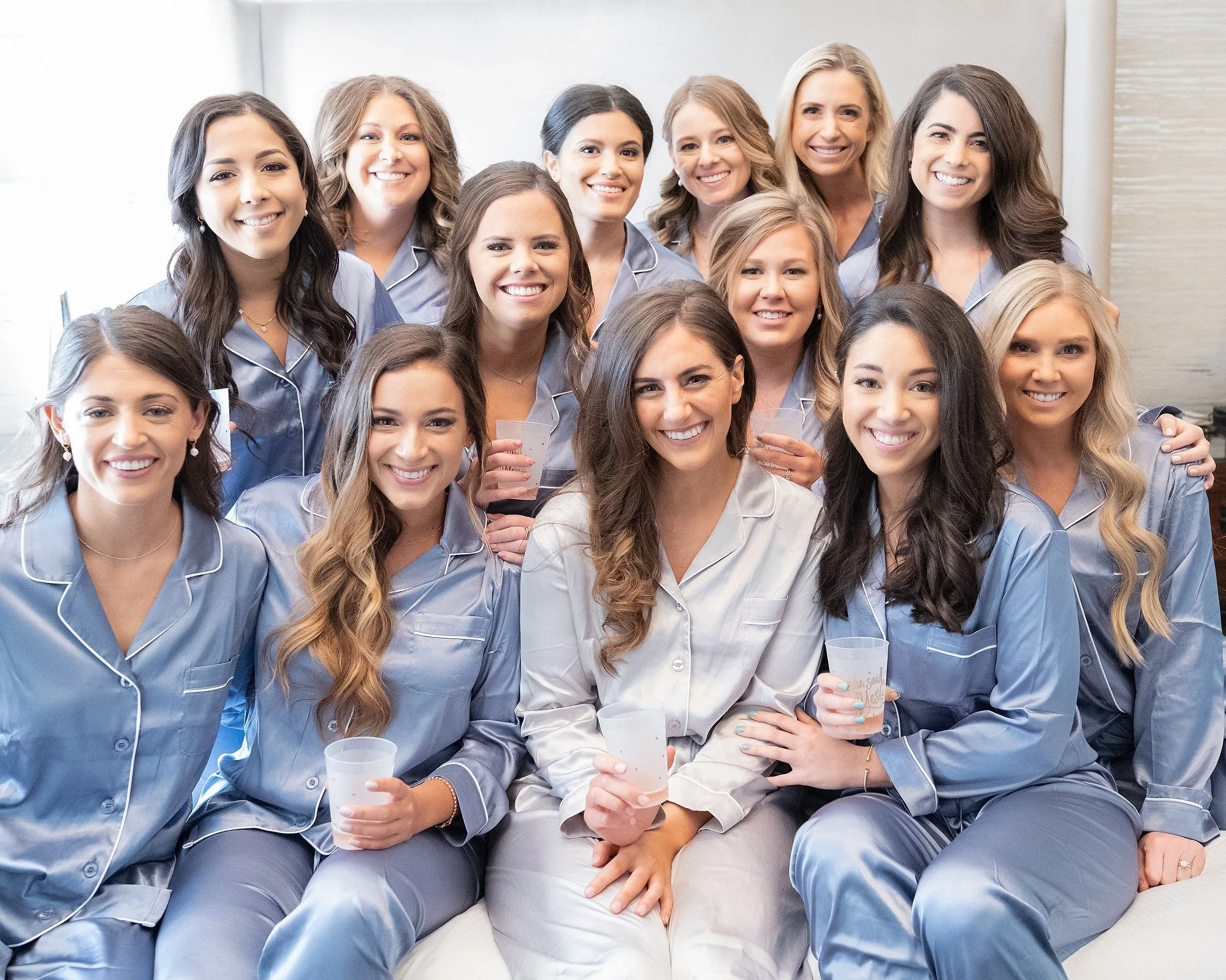 Group of women in matching pajamas smiling and holding drinks, gathered together in a cozy setting.