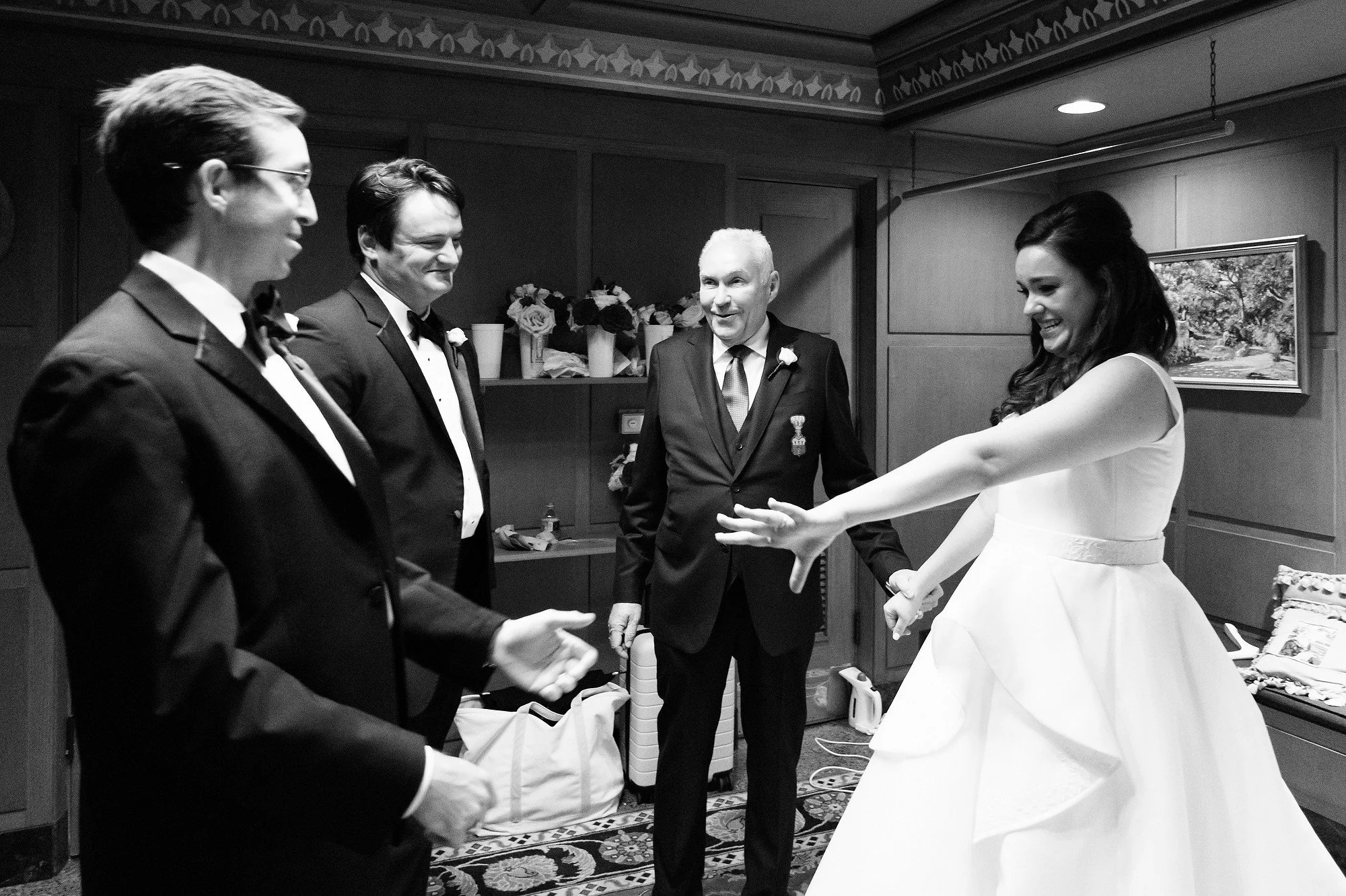 A black and white photo of a bride in a wedding dress dancing with three groomsmen in tuxedos, while an older man in a suit stands nearby smiling, in a room with wooden paneling and a framed painting on the wall.