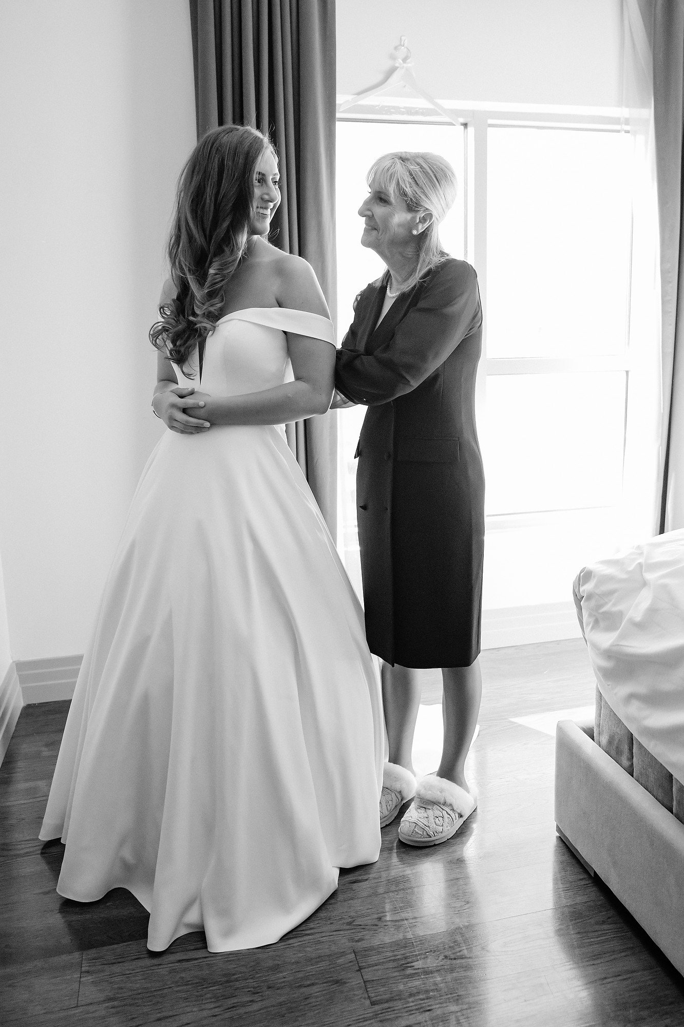 A bride in a wedding gown and an older woman in a black dress and slippers sharing a happy moment in a room with sunlight coming through the window.