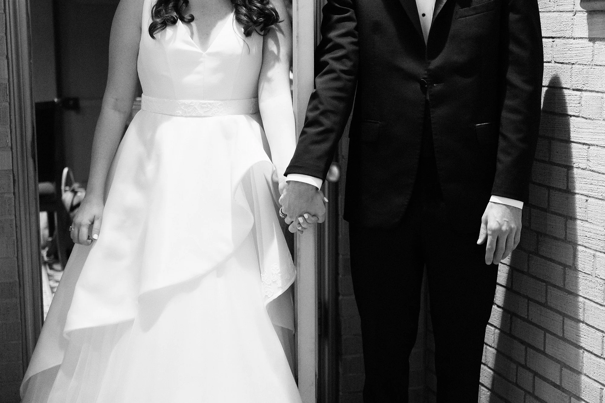 Close-up of a bride and groom holding hands during their wedding, with only the their torsos and hands visible. The bride wears a white wedding dress and the groom wears a black tuxedo.