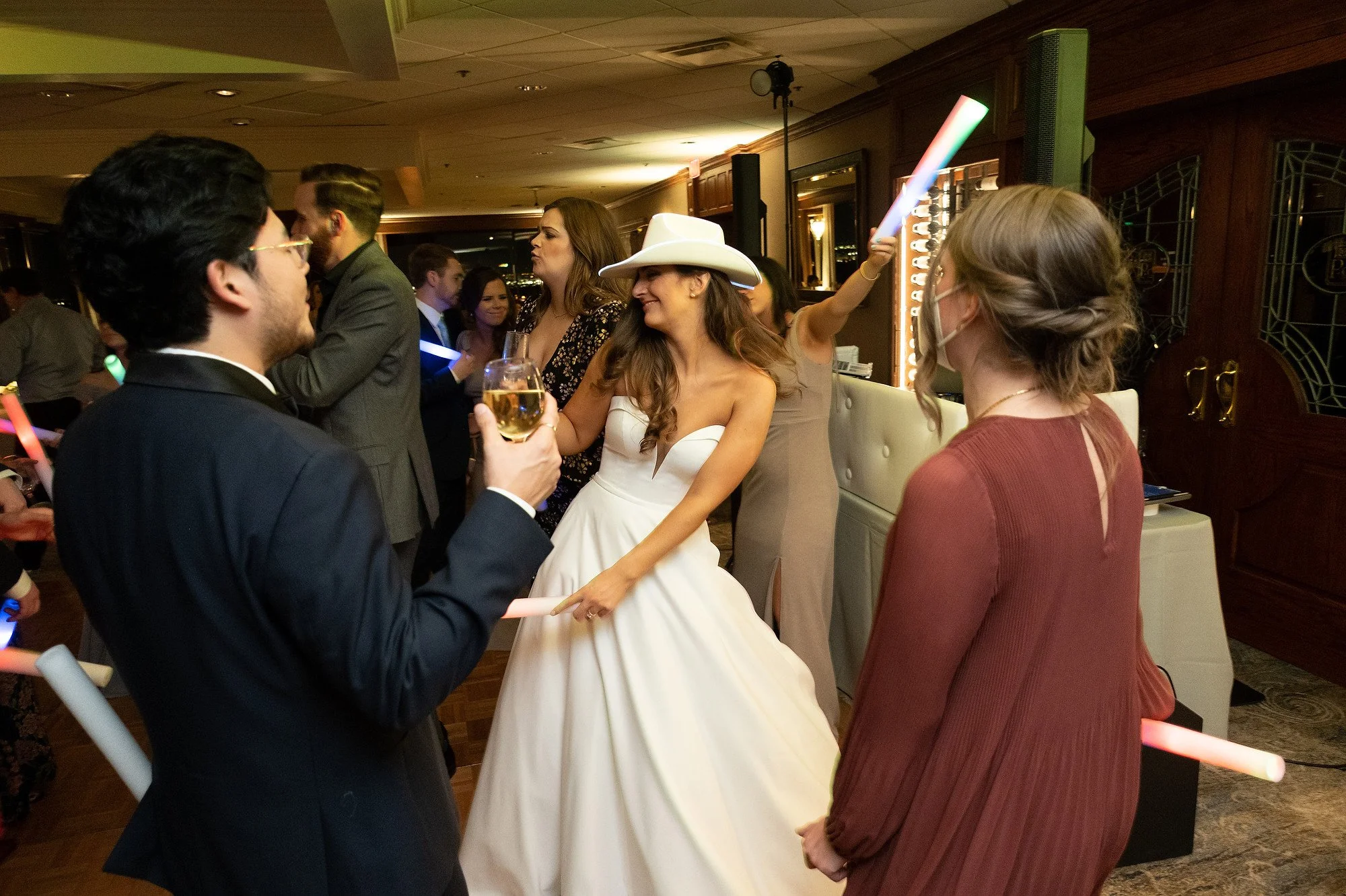 A group of people dancing and socializing at a wedding reception, with some holding glow sticks and drinks.
