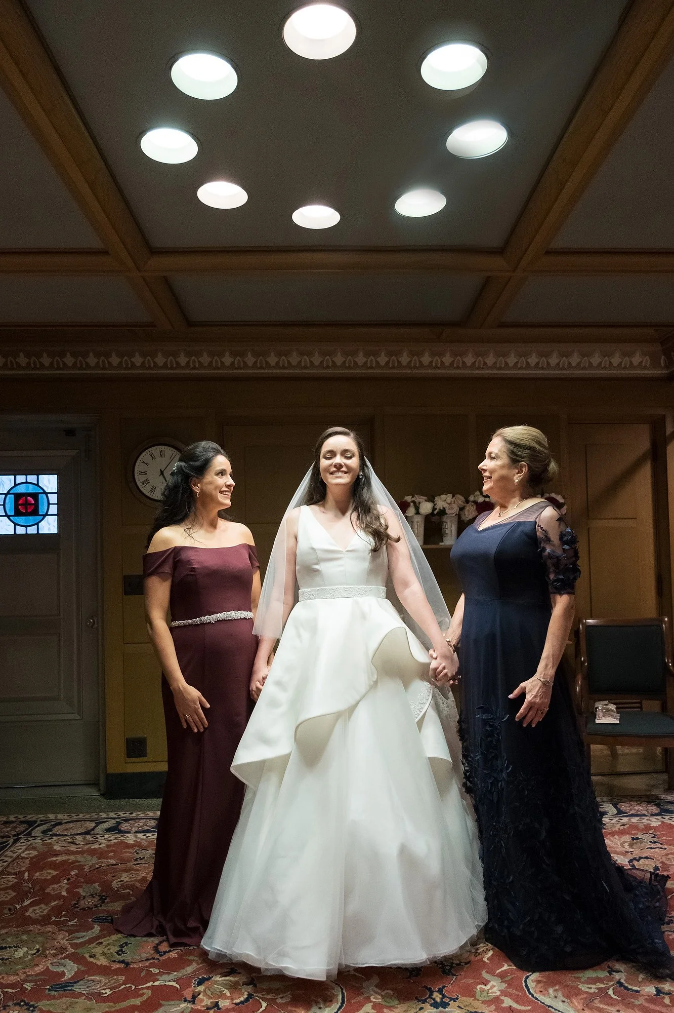 Bride holding hands with two women, dressed in formal attire, inside a wood-paneled room with a patterned carpet and a clock on the wall.