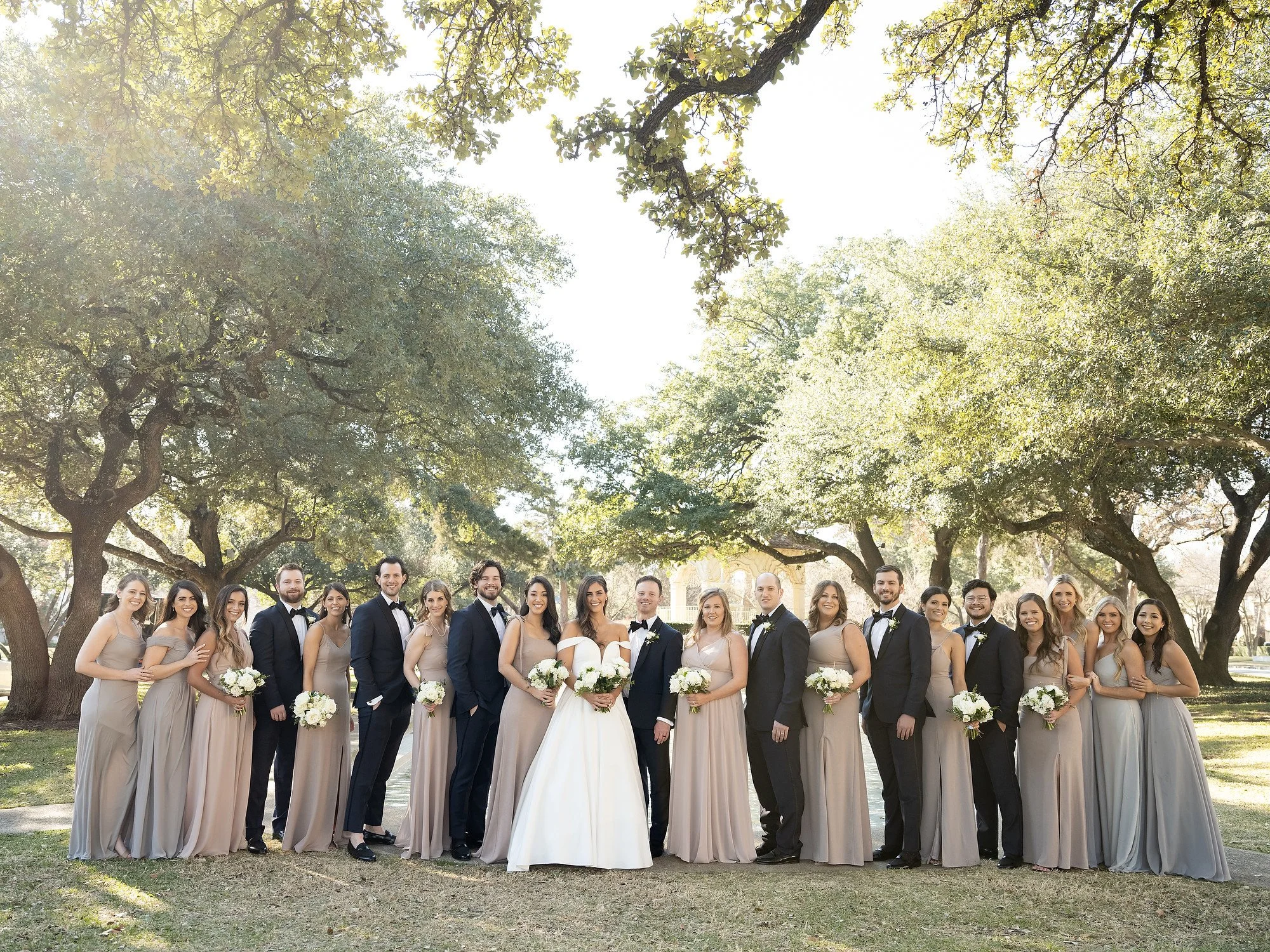 Group of wedding party members, including bride, groom, bridesmaids, and groomsmen, standing outdoors under large trees on a sunny day.