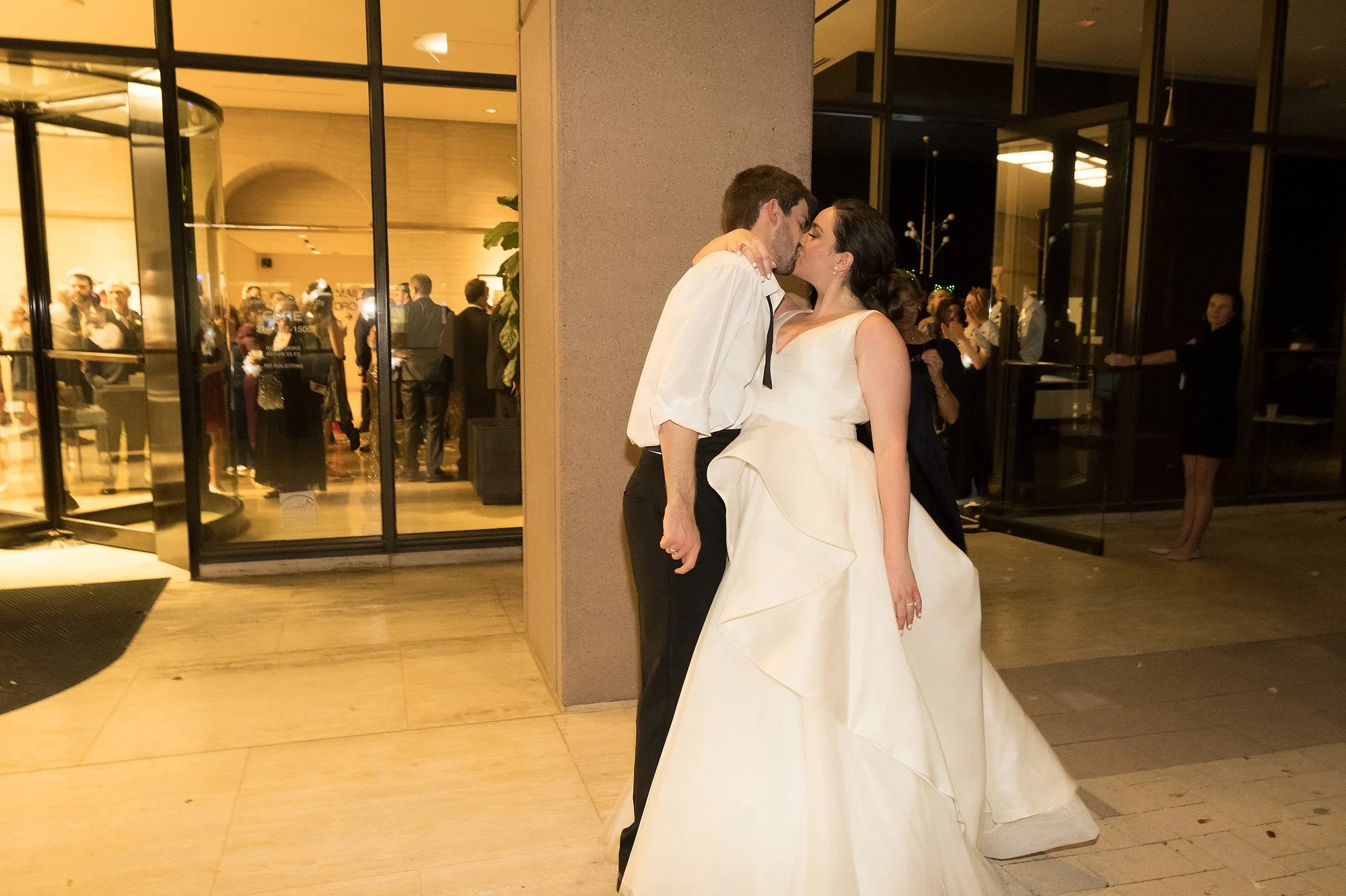 A bride and groom share a kiss outside a reception venue, with guests visible through glass doors in the background.