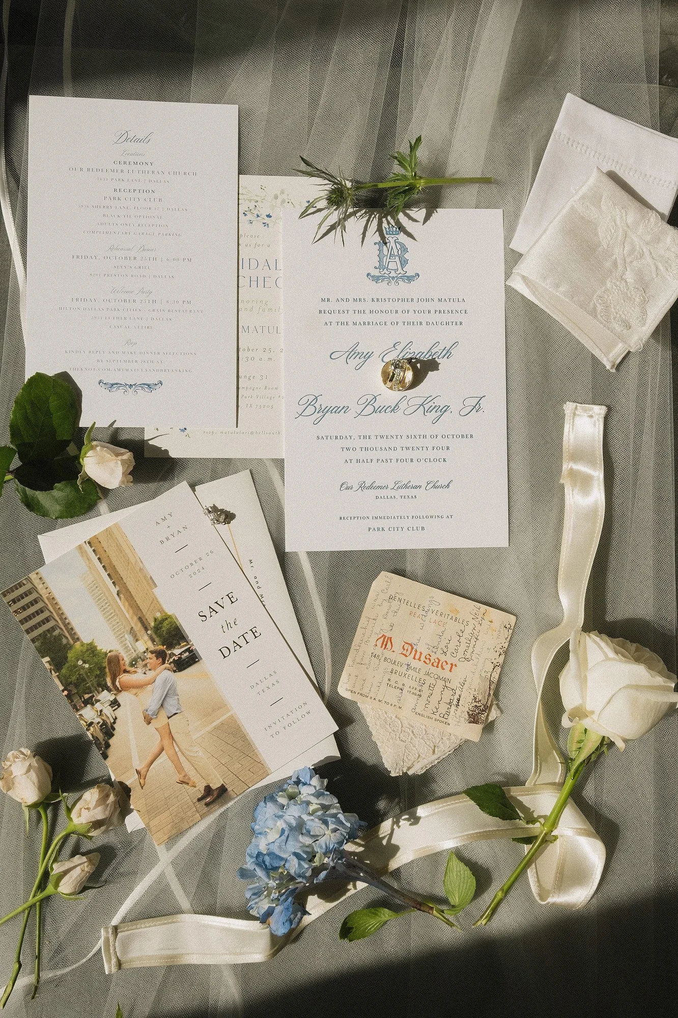 Wedding invitation suite with wedding details, a save the date card featuring a couple walking in an urban setting, vintage postcards, white roses, blue hydrangea, a crystal ring, and decorative ribbons on a gray tablecloth.