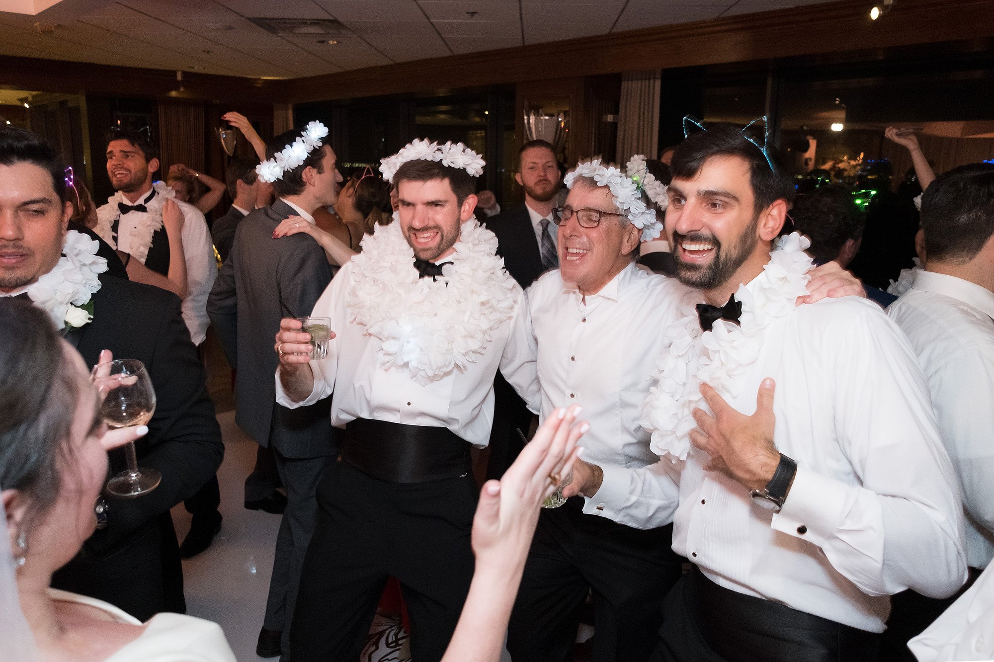 Group of men at a celebration, wearing white shirts, floral leis, and accessories, dancing and smiling at a party or wedding reception.