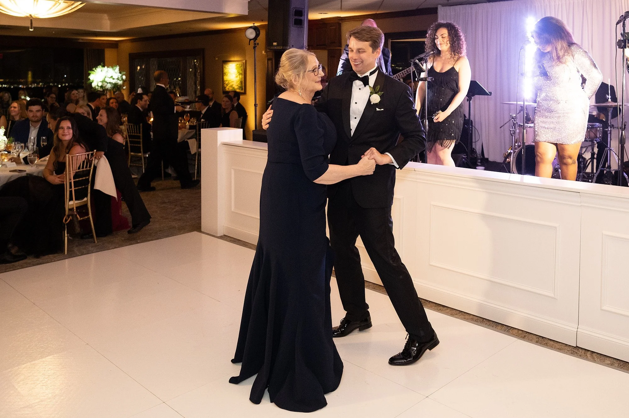 A young man in a tuxedo dances with an older woman in a navy dress at a formal event, with a band playing on stage in the background and guests seated at tables.