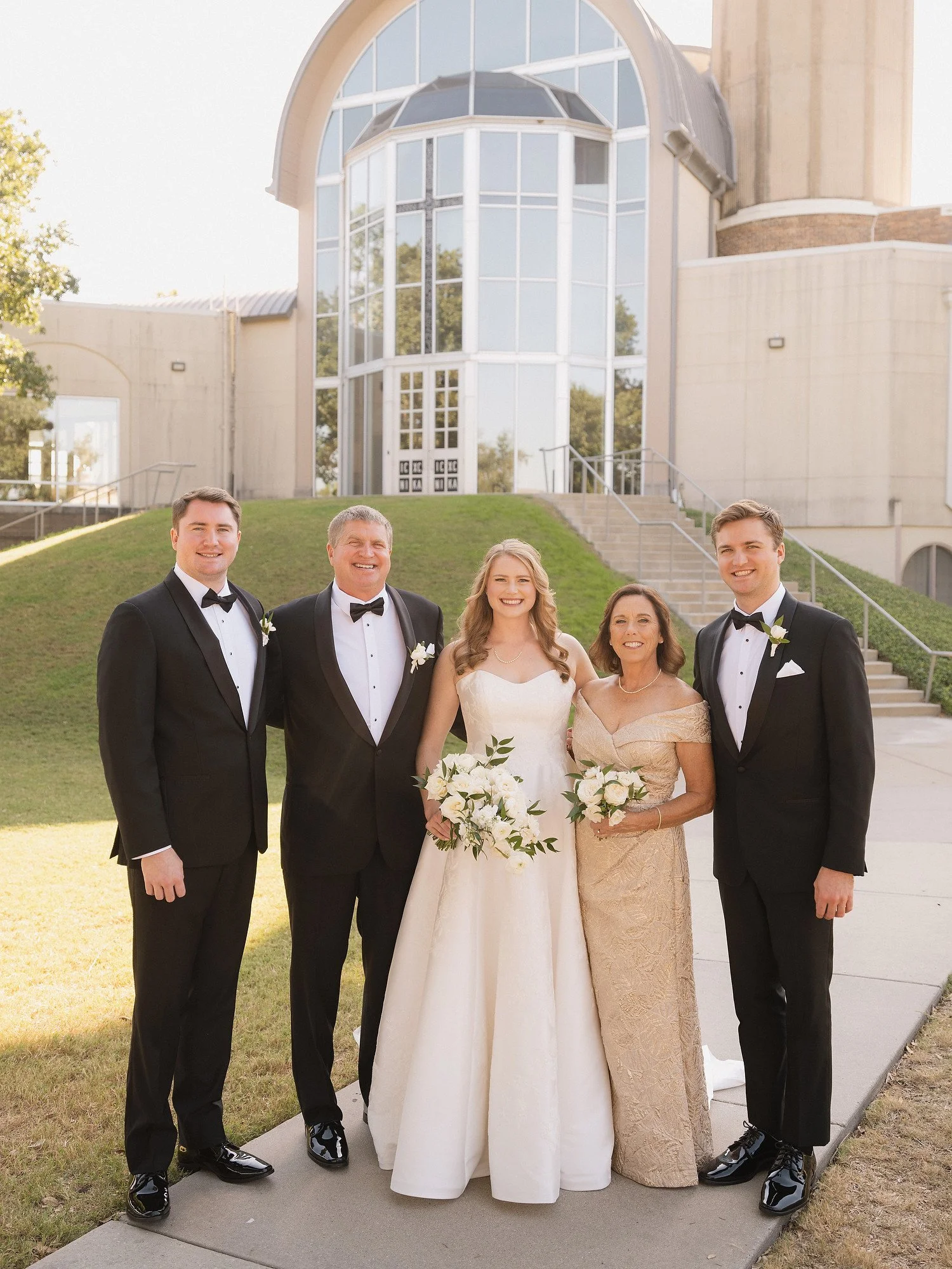 Group of five people in formal wedding attire standing outdoors on sidewalk, in front of a modern church building with large glass windows, with a grassy hill and stairs in the background.