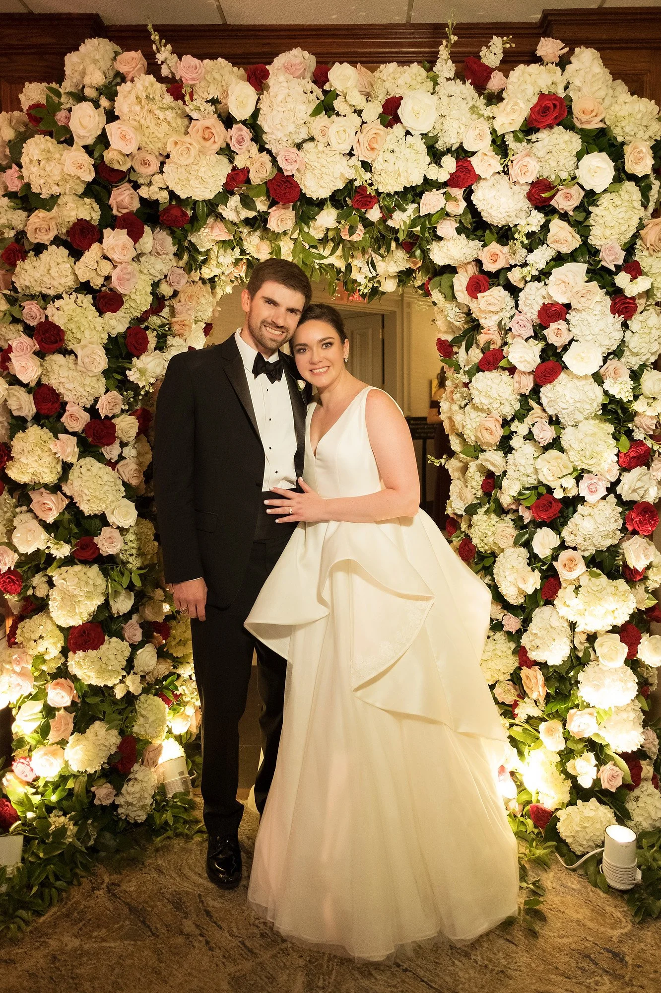 A newlywed couple smiling and standing close together, posing for a photo at their wedding, in front of a large arch made of white, pink, and red flowers with green leaves, inside an elegant venue.