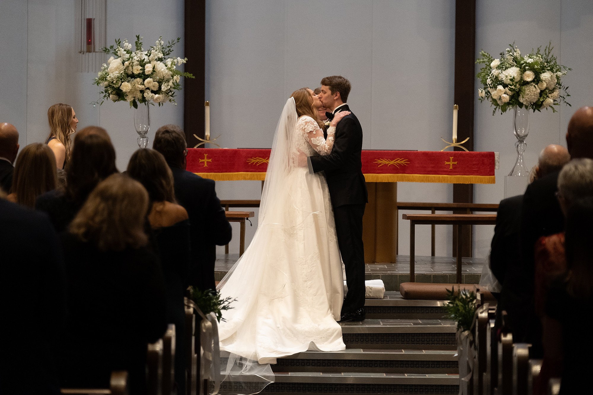 A bride and groom share a kiss at their wedding ceremony in a church, with an altar decorated with large floral arrangements and people seated in the pews watching.