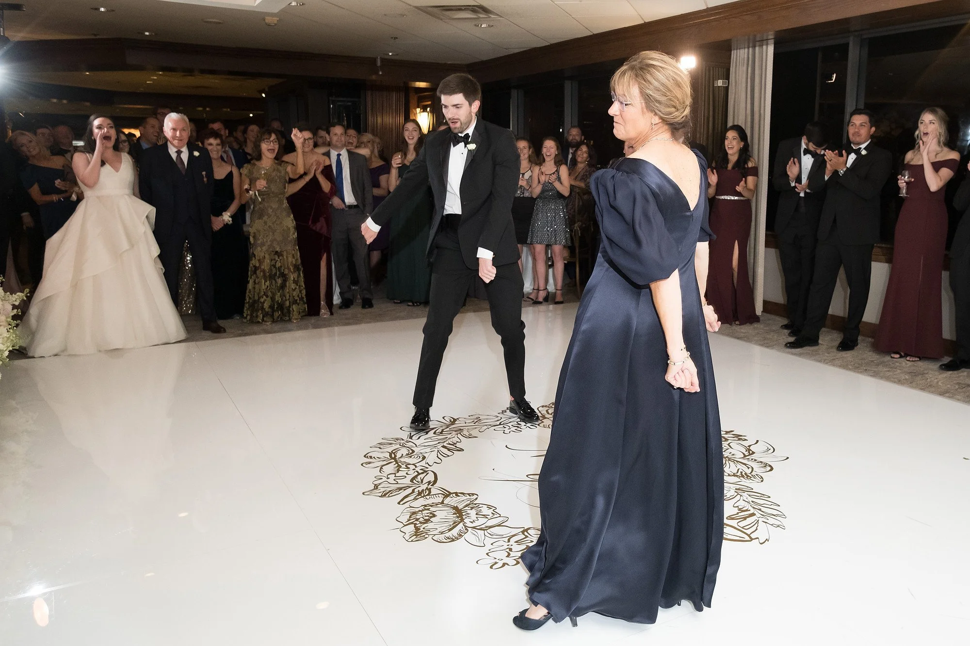 A man in a tuxedo dances with a woman in a navy blue gown on a decorated dance floor, surrounded by a crowd of people watching and cheering at a formal event.