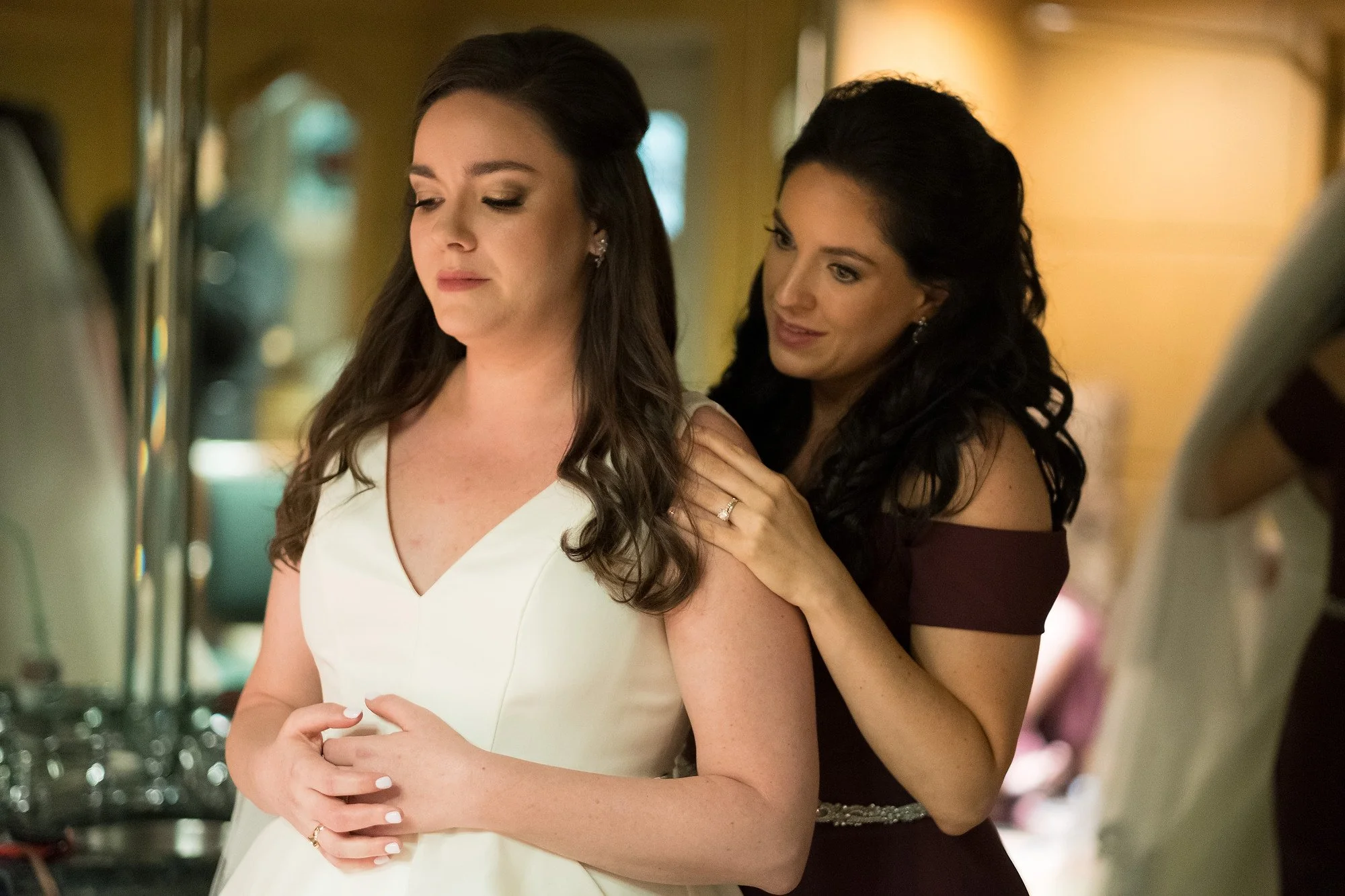 Two women, one in a white dress and the other in a dark dress, preparing for a wedding or event in an indoor setting