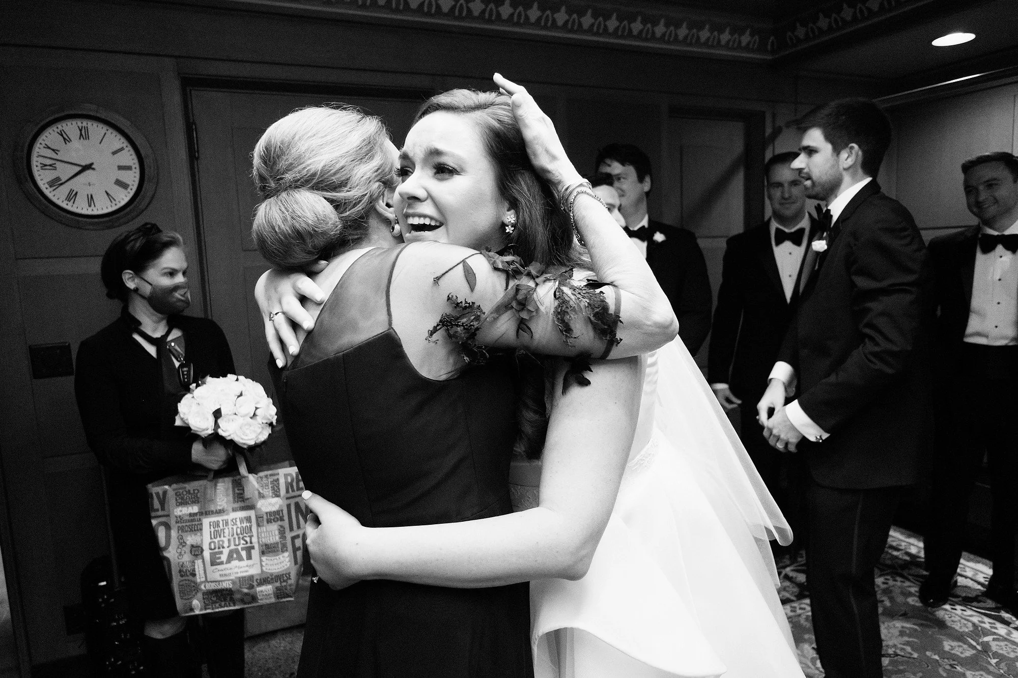 A woman in a wedding dress hugging an older woman, who is wearing a dark dress with lace sleeves, in a room with wedding party members in tuxedos and a woman holding a bouquet in the background.