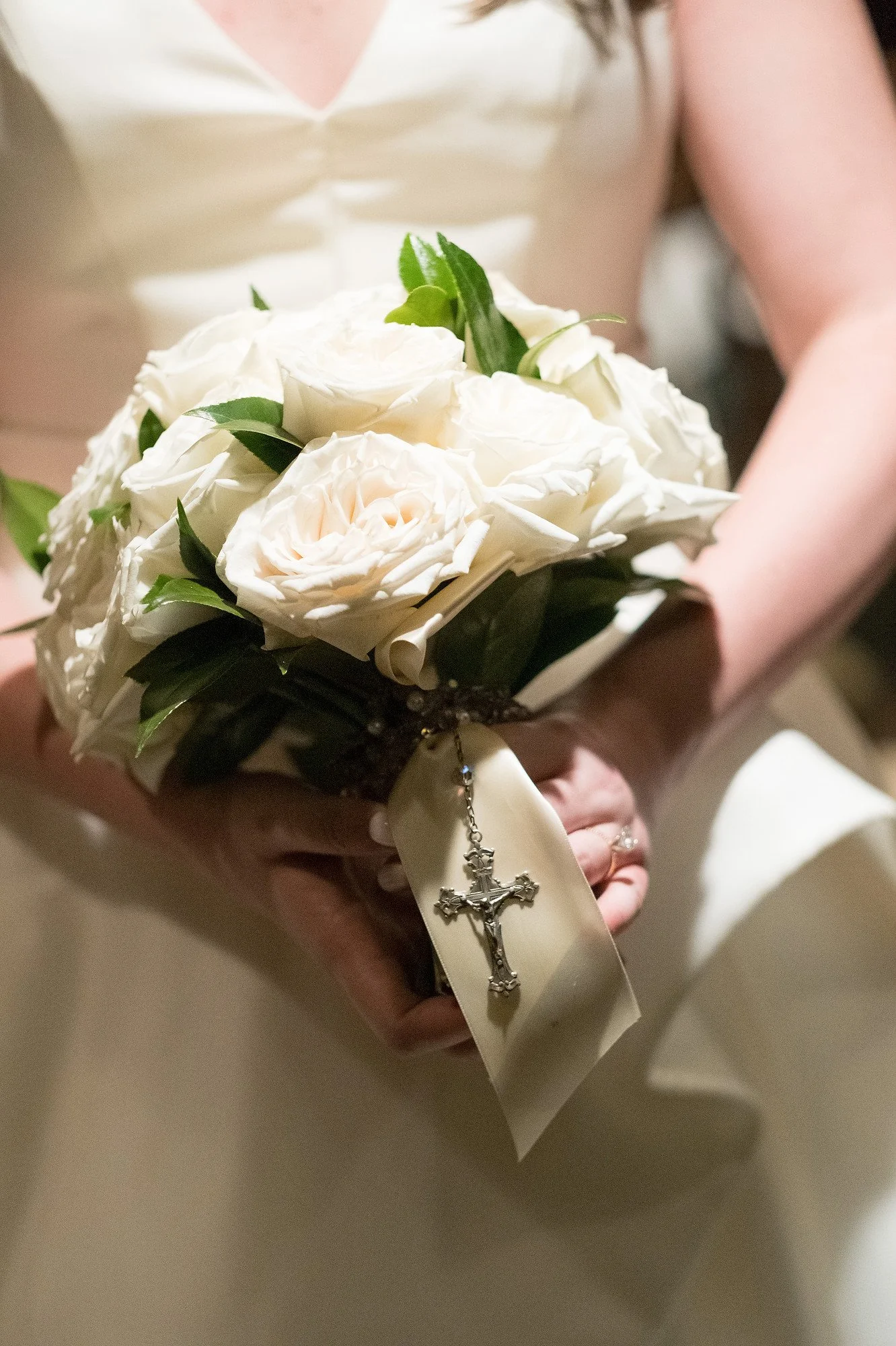 A person holding a bouquet of white roses with greenery, decorated with a beige ribbon and a silver rosary cross charm.