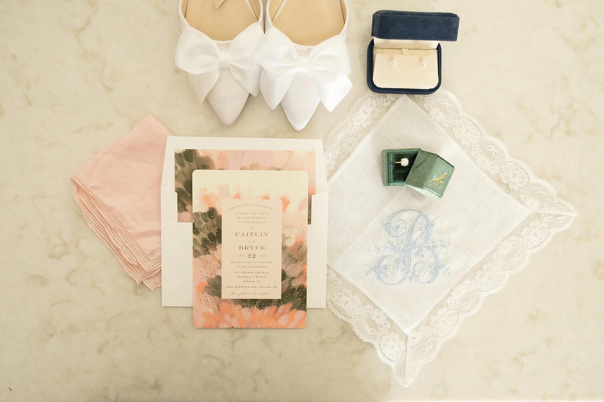 Wedding invitation, white shoes with bows, jewelry box with earrings, pink napkins, embroidered handkerchief with initials, and a ring in a green box.