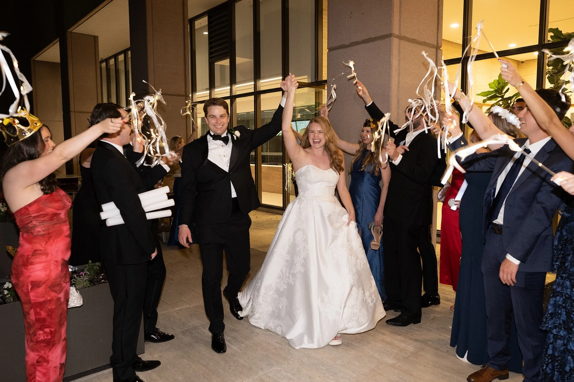 Bride and groom celebrating with wedding guests who are throwing streamers while smiling and holding celebratory items outside a modern building at night.