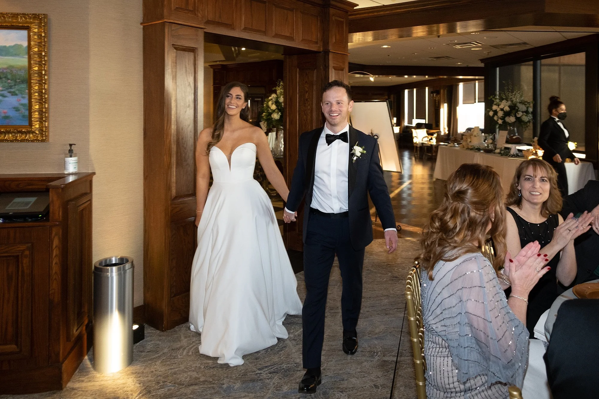 Bride and groom holding hands and smiling as they walk into a wedding reception, with guests clapping and celebrating.