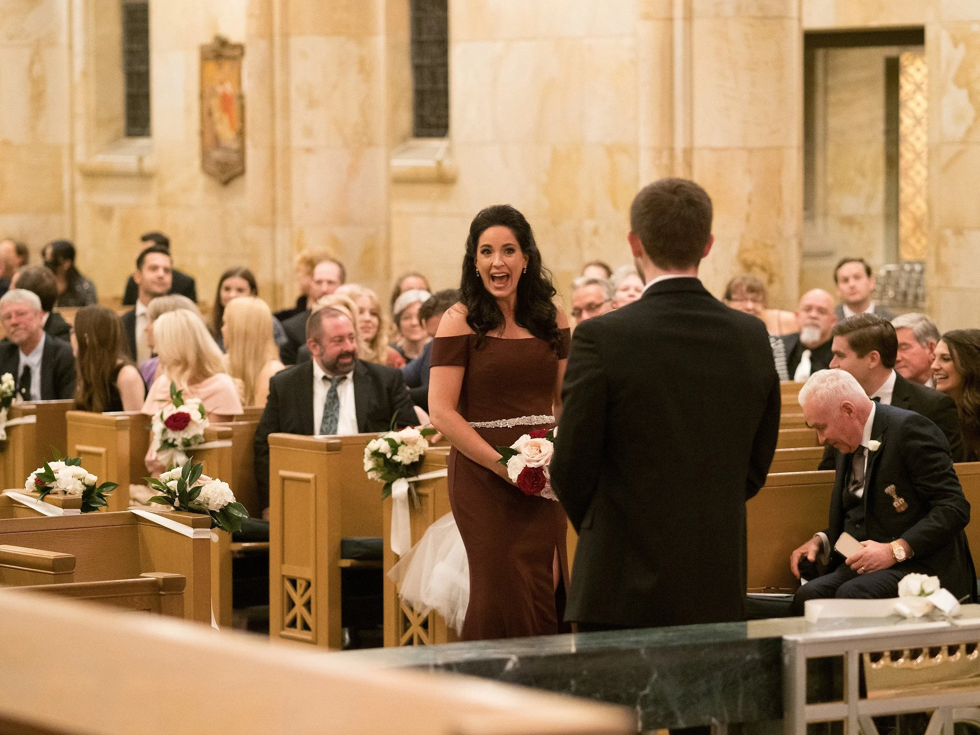 Woman in a brown dress with off-shoulder sleeves holding a bouquet of flowers, smiling and talking to a man in a black suit during a wedding ceremony inside a church filled with seated guests.